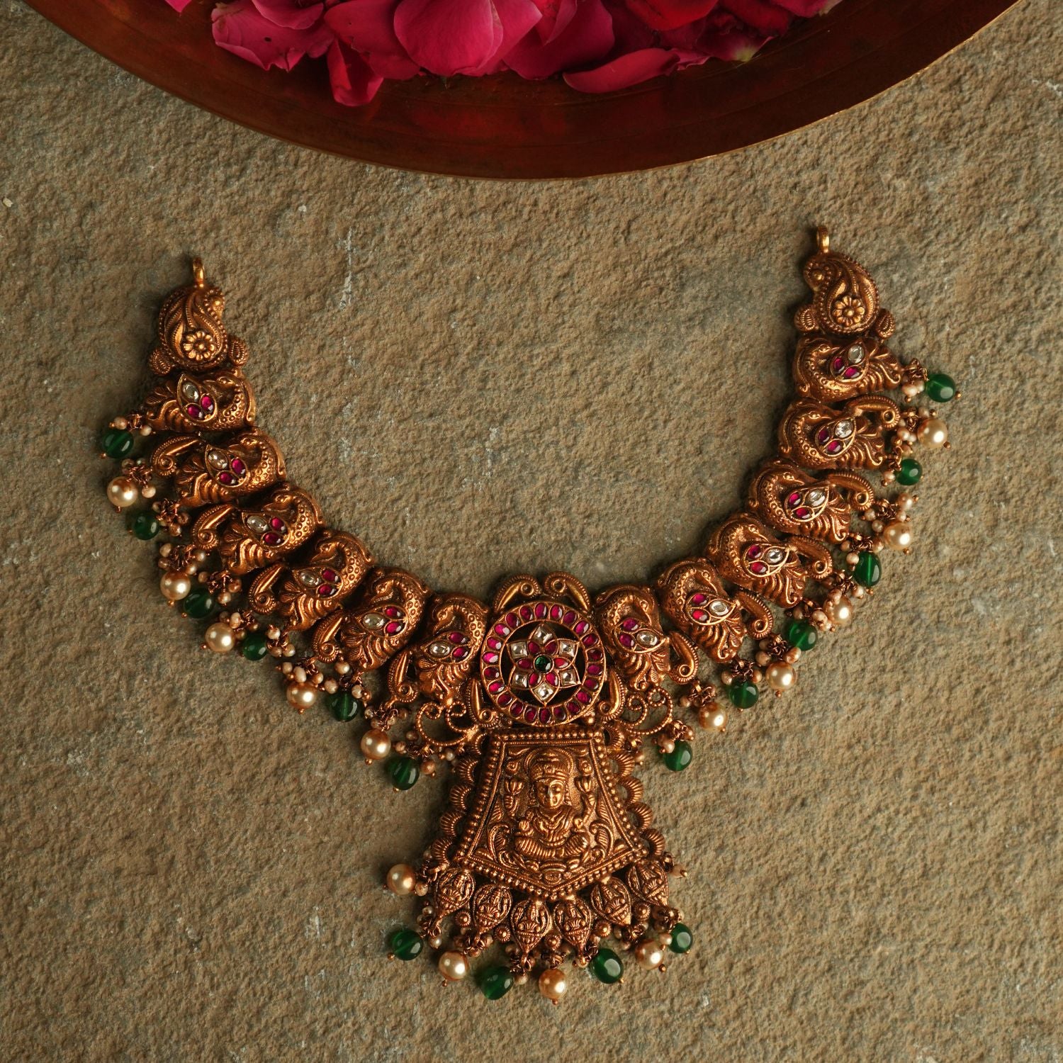 An antique gold-plated silver necklace adorned with peacock and goddess motifs radiates timeless splendor, enriched with red, white, and green kundan stones, pearl droplets, and green beads.