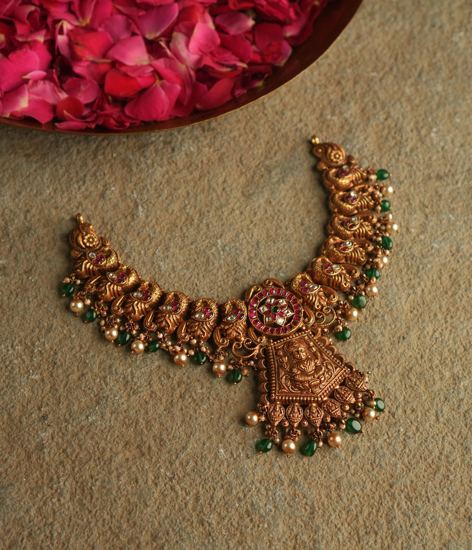 An antique gold-plated silver necklace adorned with peacock and goddess motifs radiates timeless splendor, enriched with red, white, and green kundan stones, pearl droplets, and green beads.