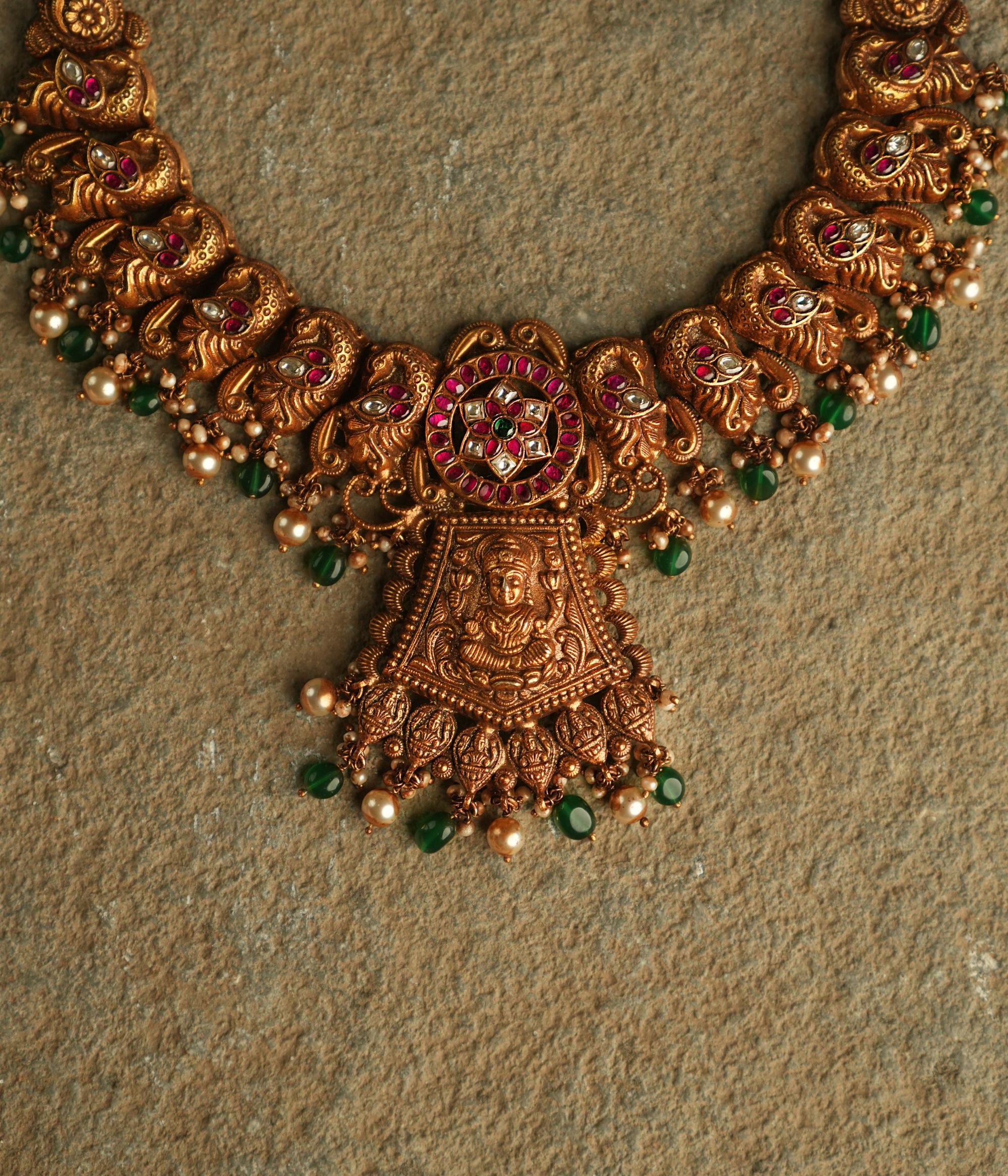 An antique gold-plated silver necklace adorned with peacock and goddess motifs radiates timeless splendor, enriched with red, white, and green kundan stones, pearl droplets, and green beads.