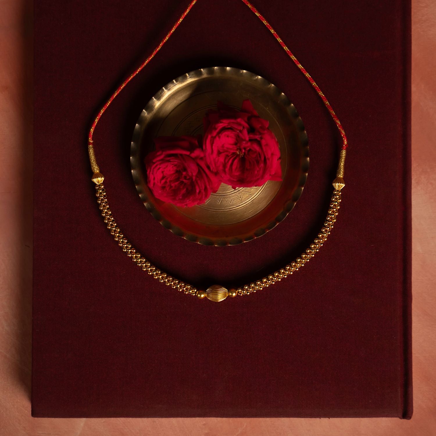 Gold-plated silver necklace featuring a delicate arrangement of small gold beads with a larger, textured gold bead at the center. Displayed on deep red fabric, accompanied by a brass bowl and red roses in the background, exuding elegance and traditional charm.