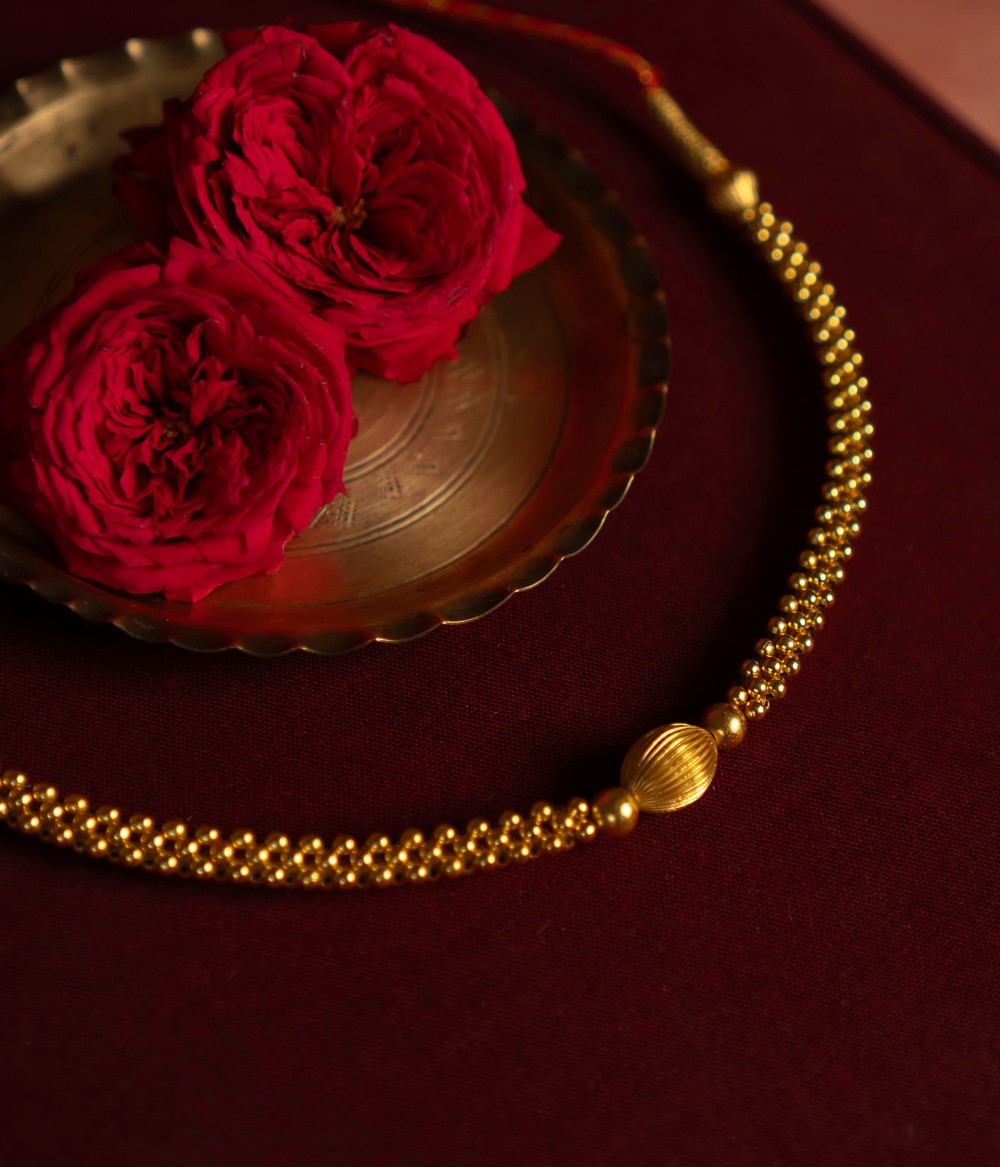 Gold-plated silver necklace featuring a delicate arrangement of small gold beads with a larger, textured gold bead at the center. Displayed on deep red fabric, accompanied by a brass bowl and red roses in the background, exuding elegance and traditional charm.