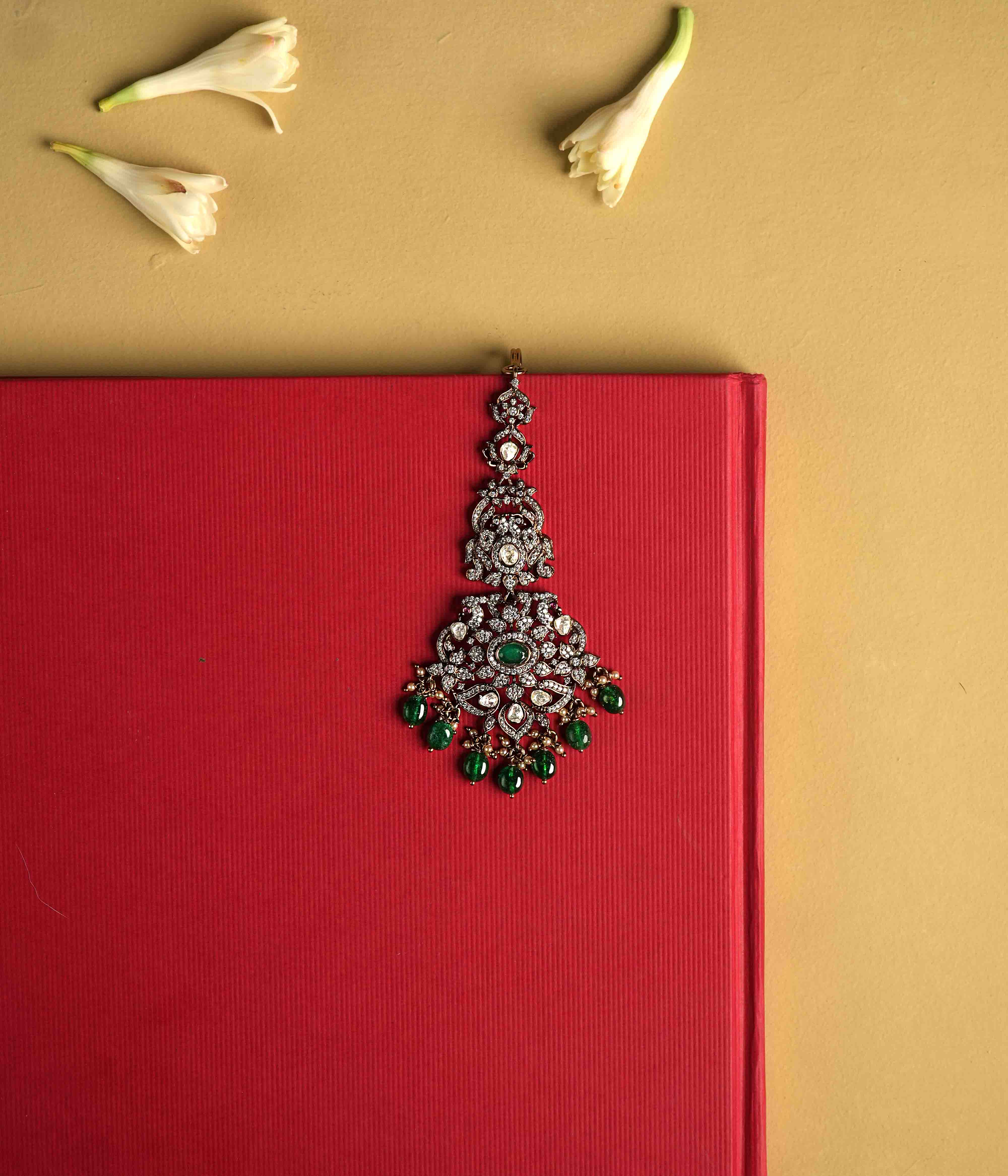 This intricately crafted Maangtika, handmade from 92.5 sterling silver with 14ct dark antique gold plating, features polki stones, CZ stones, green gemstones, green beads, and pearls. The design shines with polki and moissanite brilliance, with cultured emerald dangles adding a touch of majestic elegance, evoking the opulence of a Victorian era.
