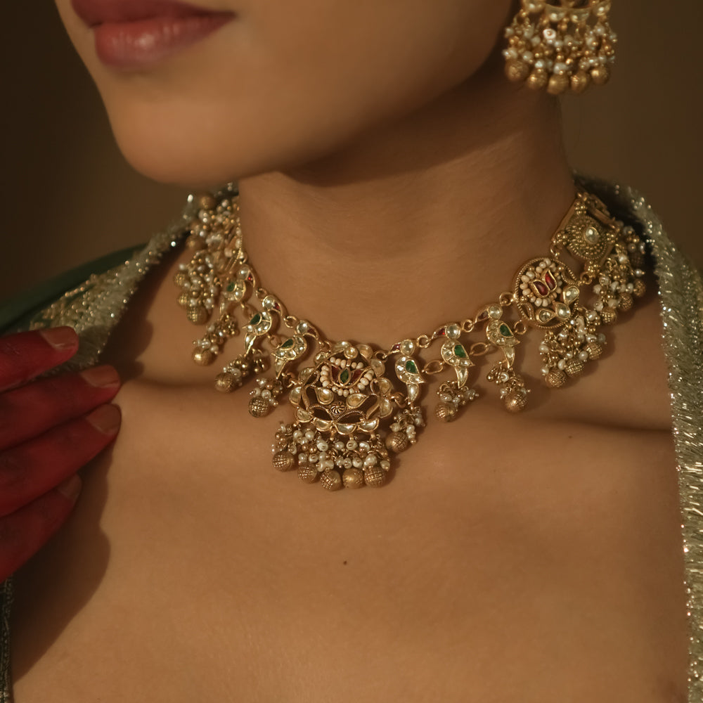 Intricate gold necklace with red and green Kundan stones, pearls, and parrot motifs.