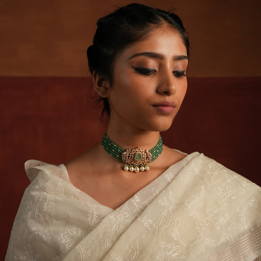 A stunning gold-plated silver choker featuring five layers of rich green beads, exuding elegance and tradition. The centerpiece showcases a bold green gemstone encased in a floral design adorned with exquisite Polki stones. Delicate pearl drops and green beads enhance its charm, making it a timeless statement piece for any occasion.