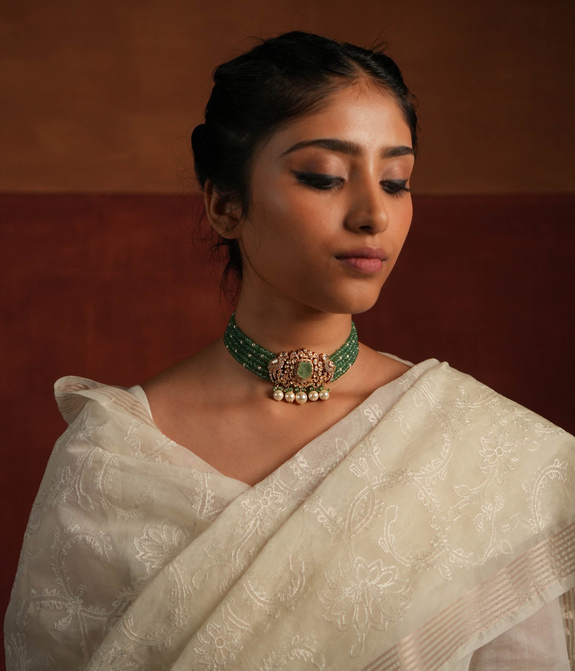 A stunning gold-plated silver choker featuring five layers of rich green beads, exuding elegance and tradition. The centerpiece showcases a bold green gemstone encased in a floral design adorned with exquisite Polki stones. Delicate pearl drops and green beads enhance its charm, making it a timeless statement piece for any occasion.