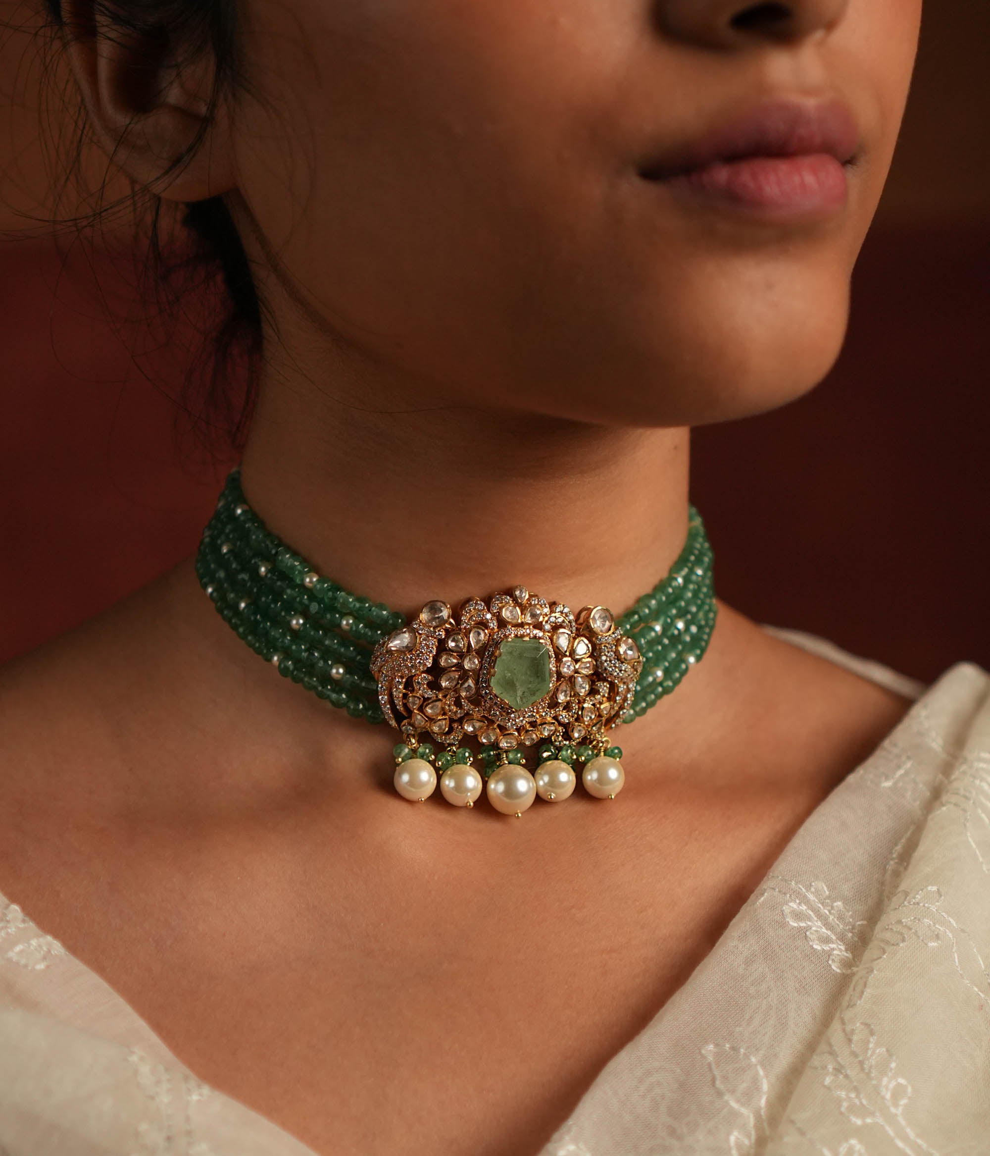 A stunning gold-plated silver choker featuring five layers of rich green beads, exuding elegance and tradition. The centerpiece showcases a bold green gemstone encased in a floral design adorned with exquisite Polki stones. Delicate pearl drops and green beads enhance its charm, making it a timeless statement piece for any occasion.