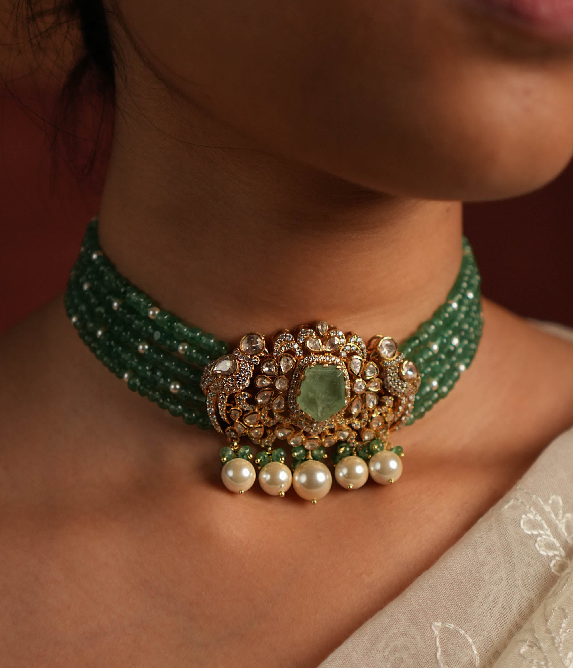 A stunning gold-plated silver choker featuring five layers of rich green beads, exuding elegance and tradition. The centerpiece showcases a bold green gemstone encased in a floral design adorned with exquisite Polki stones. Delicate pearl drops and green beads enhance its charm, making it a timeless statement piece for any occasion.