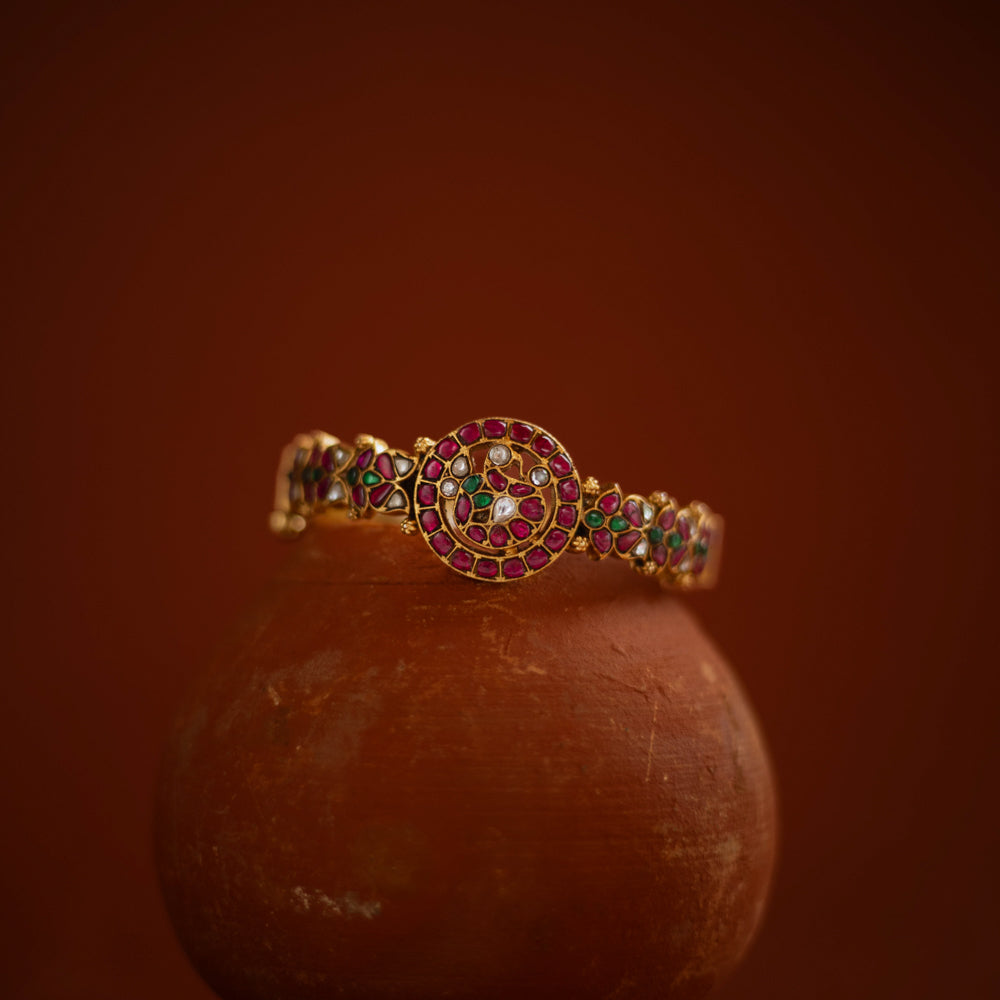 Gold bangle with circular kundan design, featuring red, green, and white stones in traditional pattern.