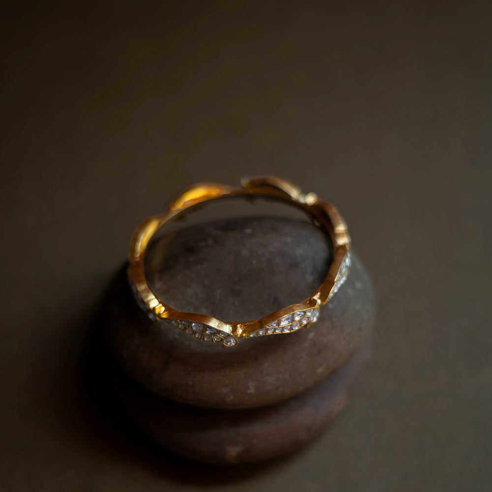 Close-up of a delicate gold bangle. Small, sparkling stones are set in a repeating, wave-like pattern along its circumference.
