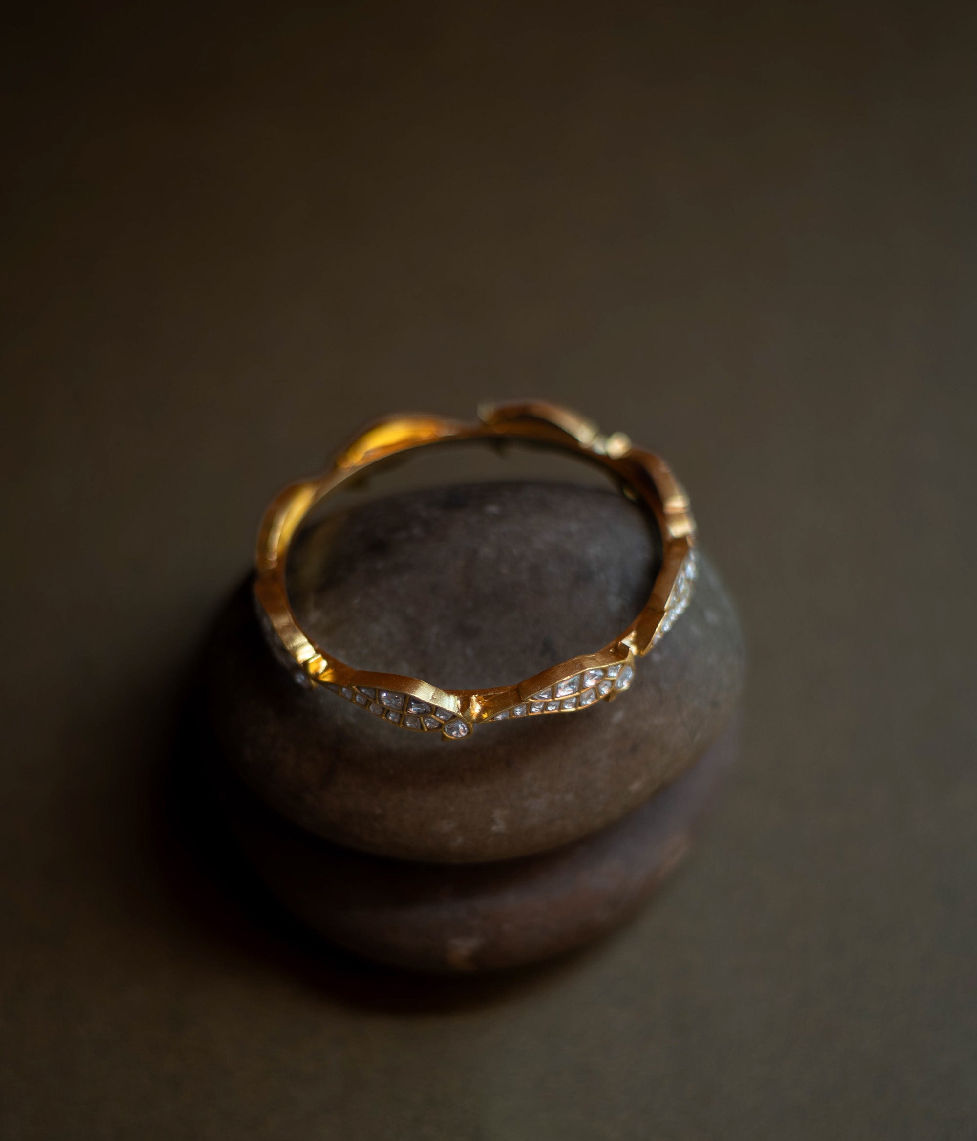 Close-up of a delicate gold bangle. Small, sparkling stones are set in a repeating, wave-like pattern along its circumference.