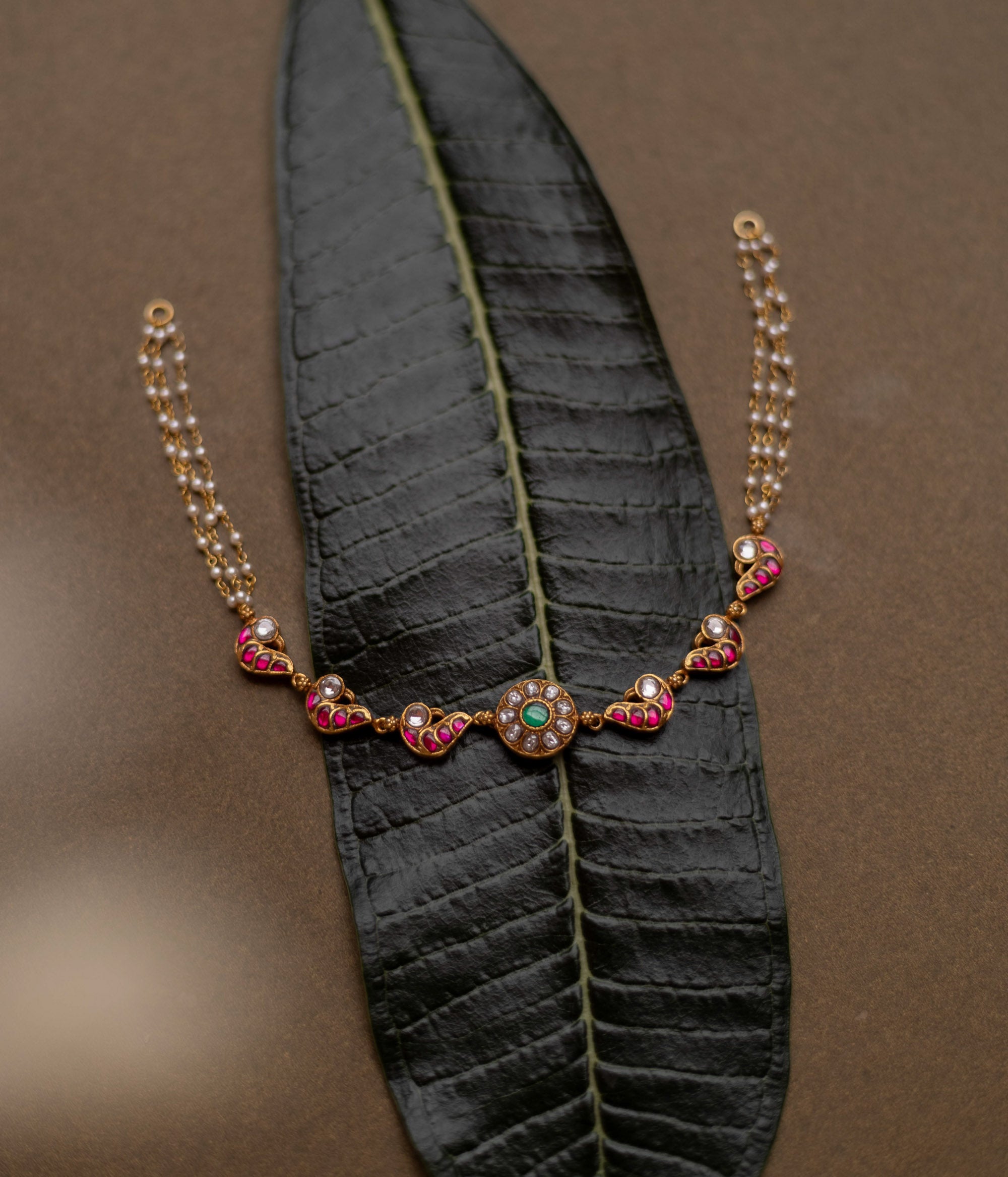 Gold-toned necklace with red, white, and green kundan stones and pearl chains, displayed on a dark green leaf.