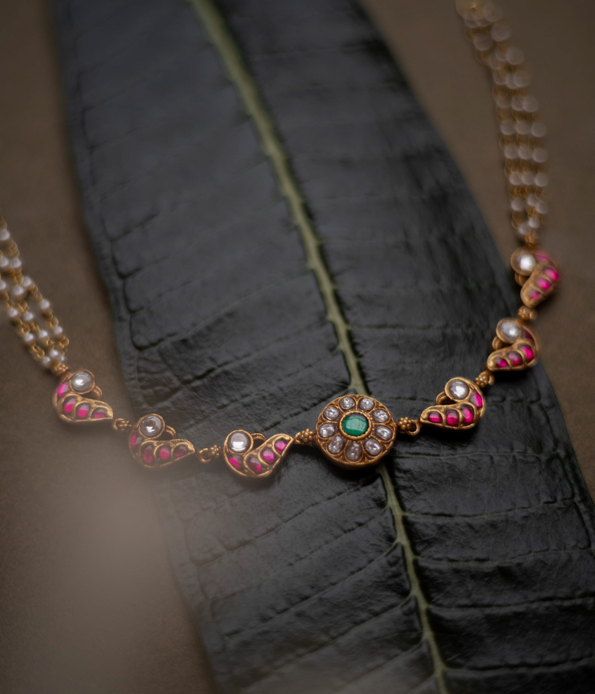 Gold-toned necklace with red, white, and green kundan stones and pearl chains, displayed on a dark green leaf.