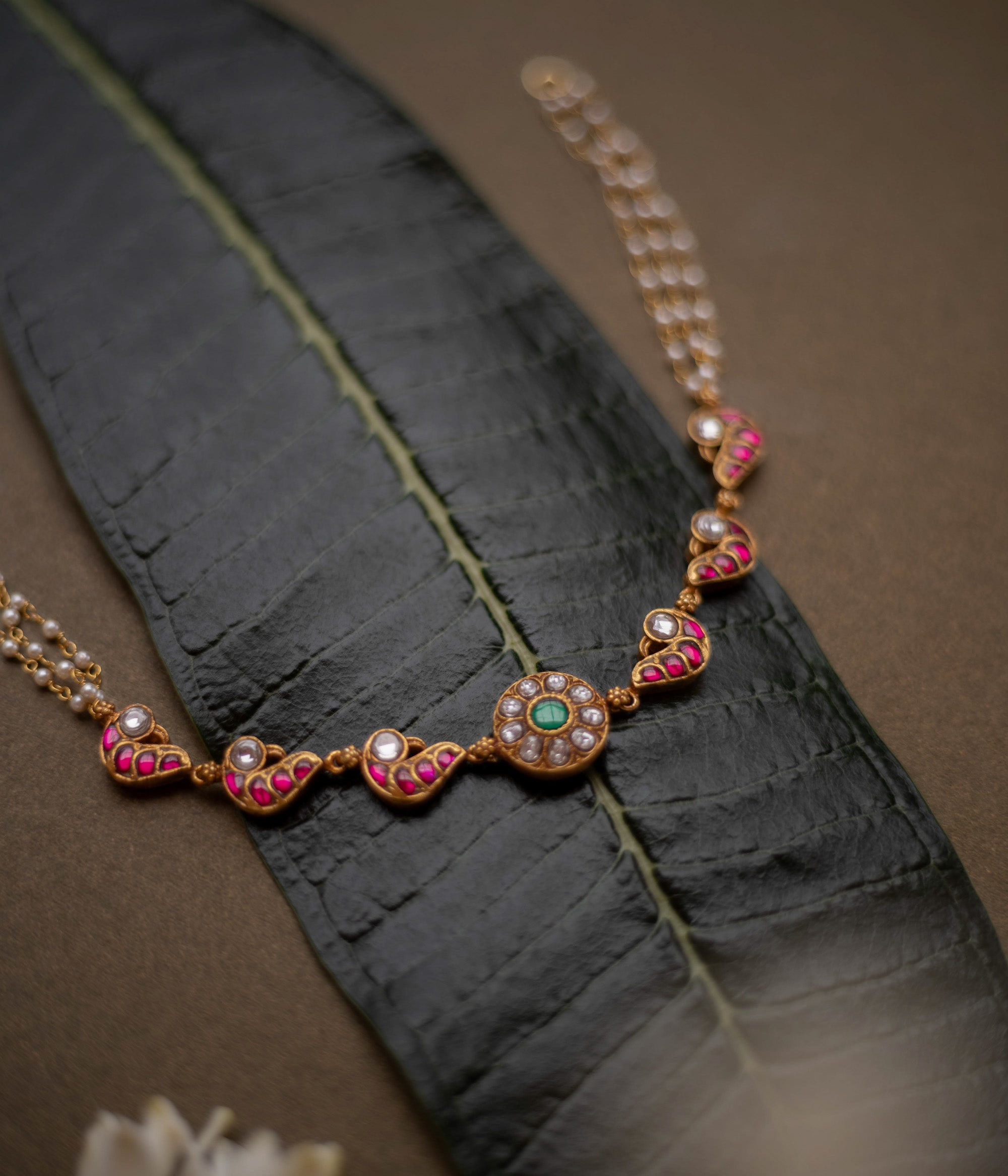 Gold-toned necklace with red, white, and green kundan stones and pearl chains, displayed on a dark green leaf.