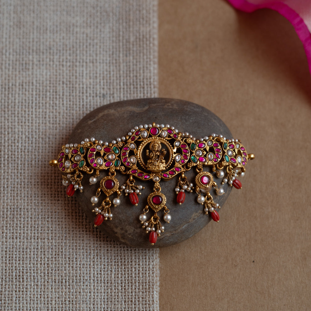 Intricate Kundan choker with a central deity. Features red, green, and white stones, red beads, and pearls.