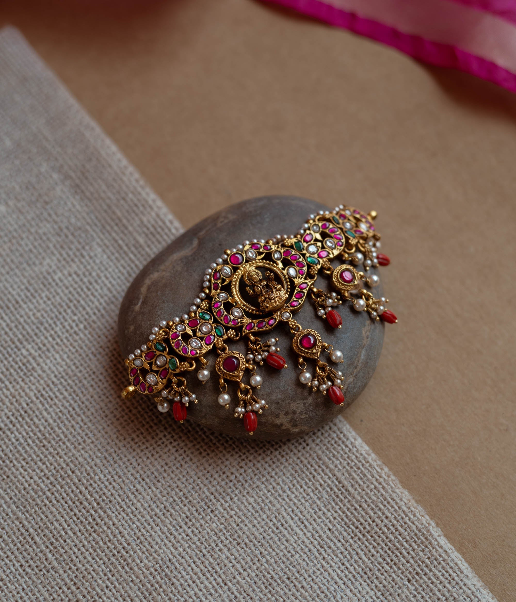 Intricate Kundan choker with a central deity. Features red, green, and white stones, red beads, and pearls.