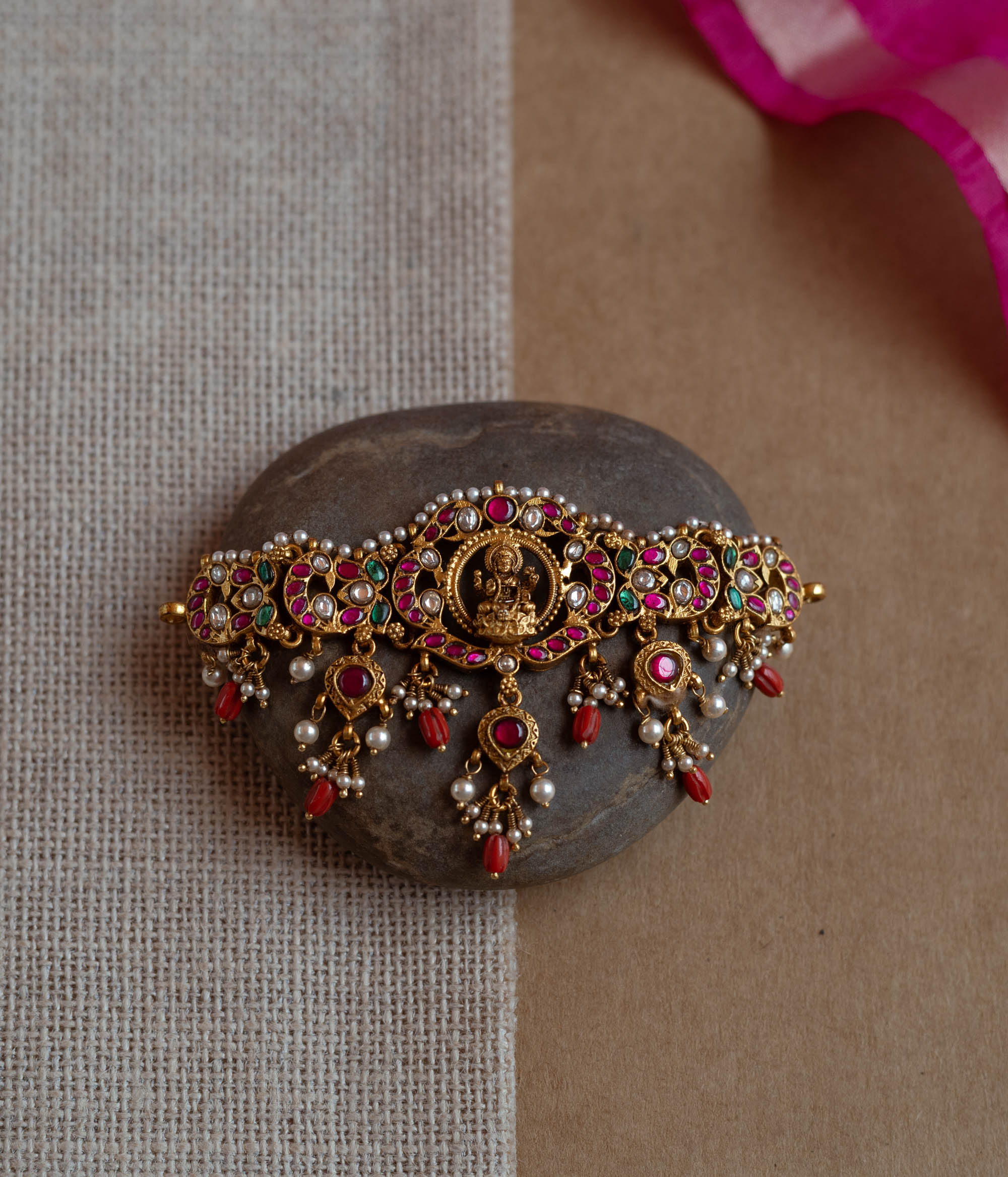 Intricate Kundan choker with a central deity. Features red, green, and white stones, red beads, and pearls.
