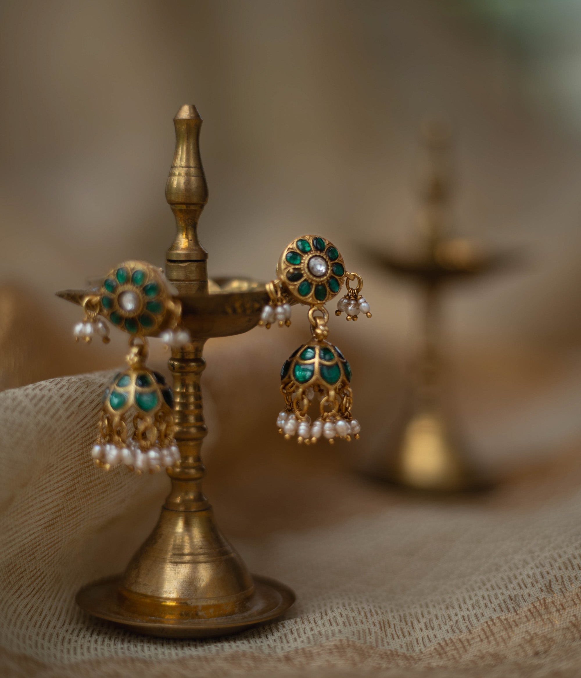 Gold jhumka earrings with green kundan stones and pearl drops, displayed on a brass oil lamp.