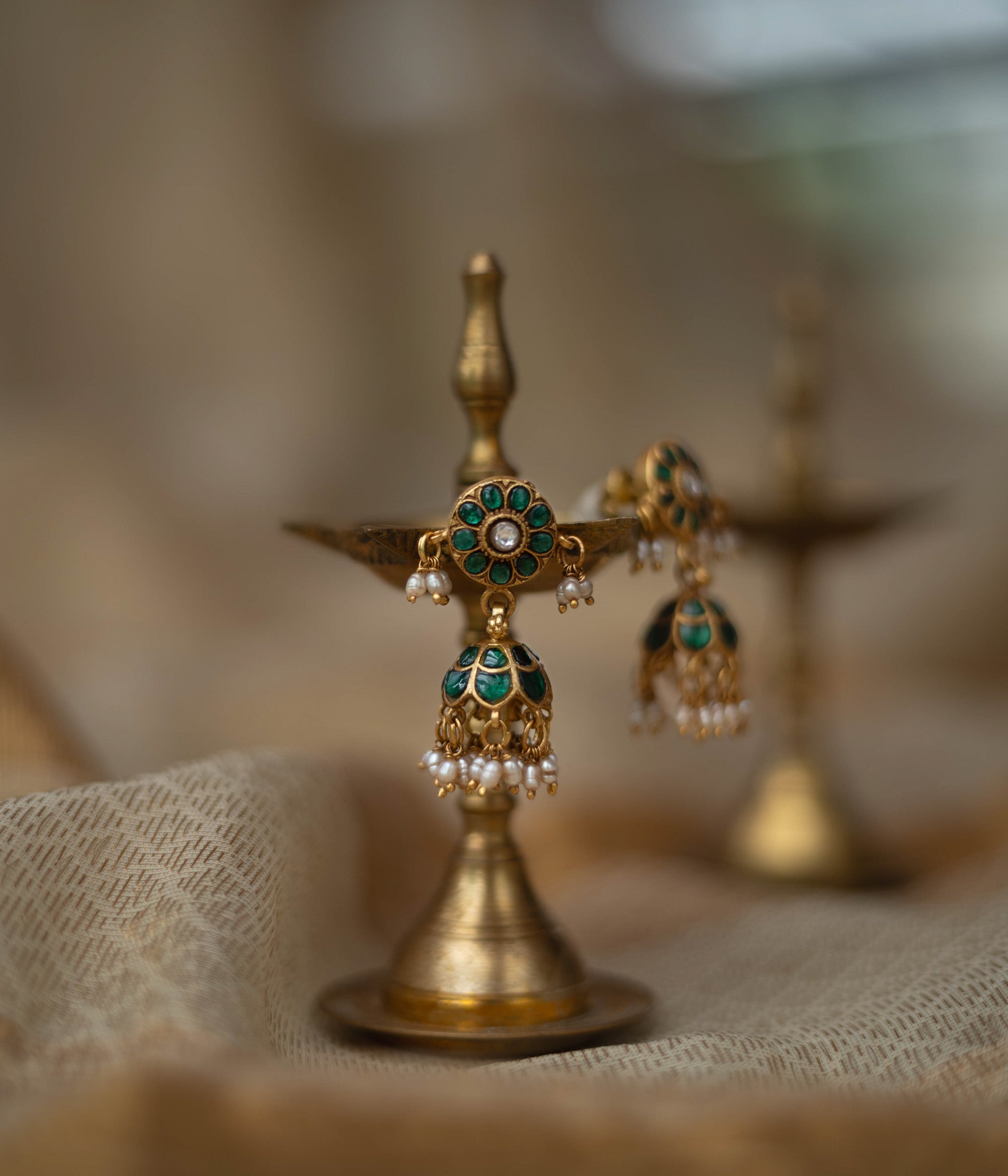 Gold jhumka earrings with green kundan stones and pearl drops, displayed on a brass oil lamp.