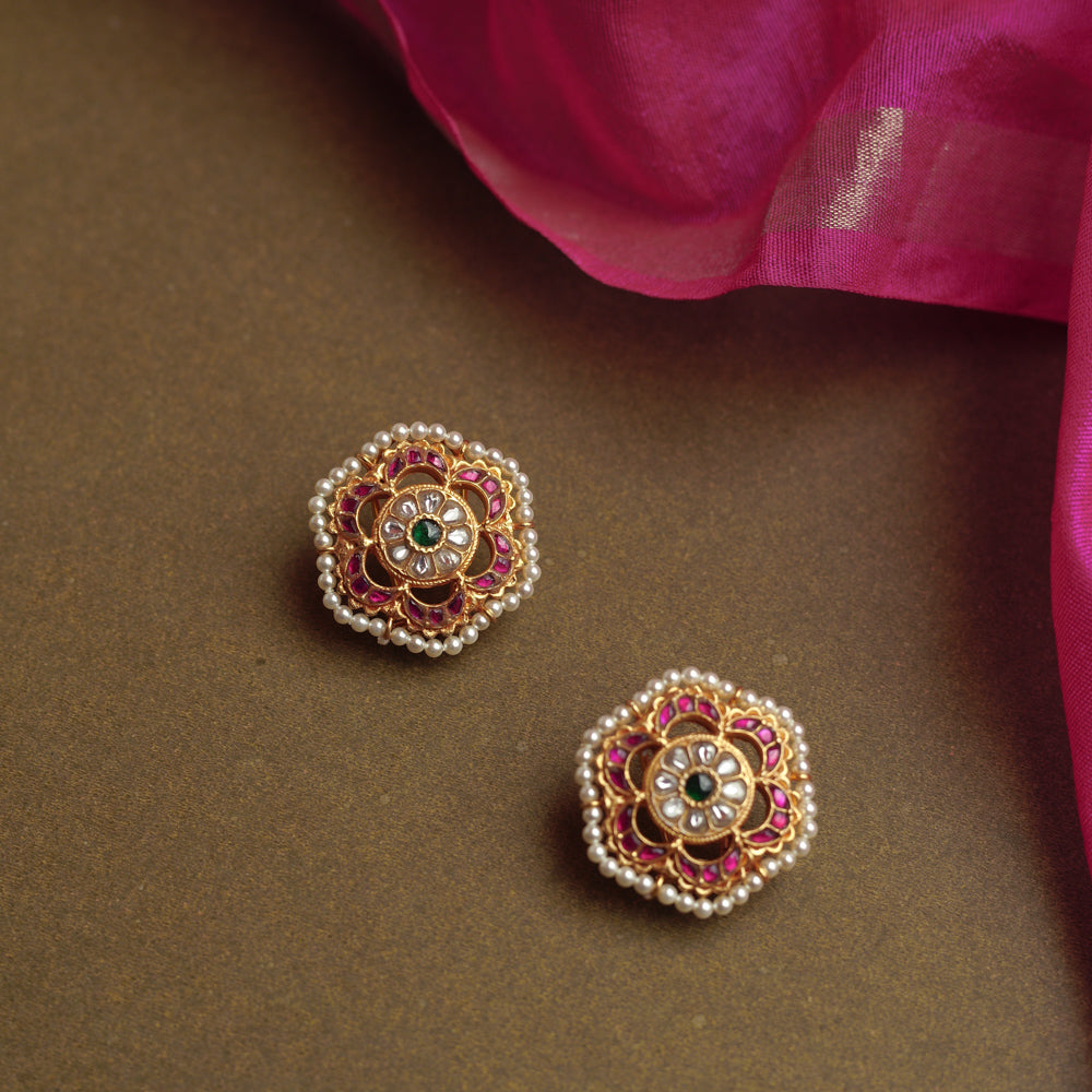 A pair of round gold stud earrings with a floral design. Each features a central green stone, surrounded by white stones, an outer ring of red stones, and a fringe of pearls.