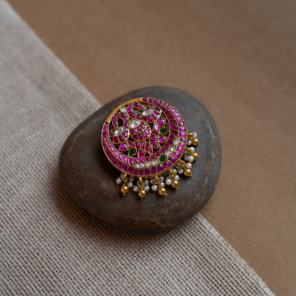 Close-up of a circular gold hairpin with a peacock motif. It's adorned with pink, green, and white Kundan stones and dangling pearls, resting on a stone.