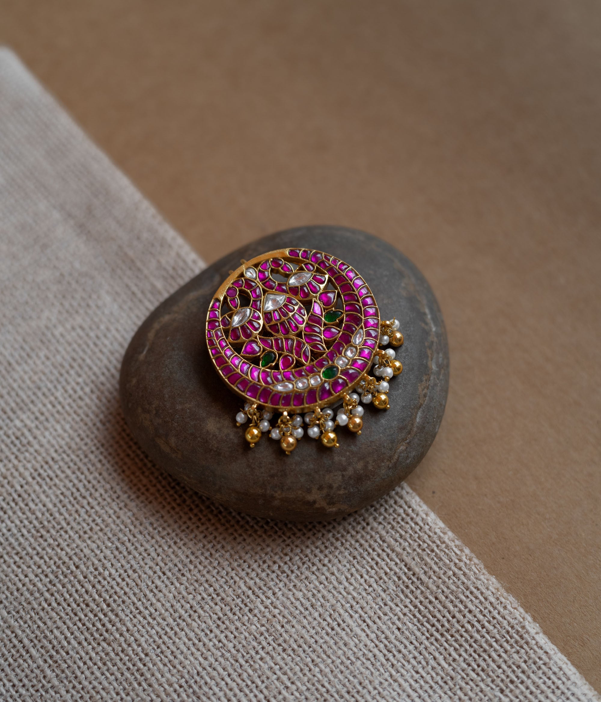 Close-up of a circular gold hairpin with a peacock motif. It's adorned with pink, green, and white Kundan stones and dangling pearls, resting on a stone.