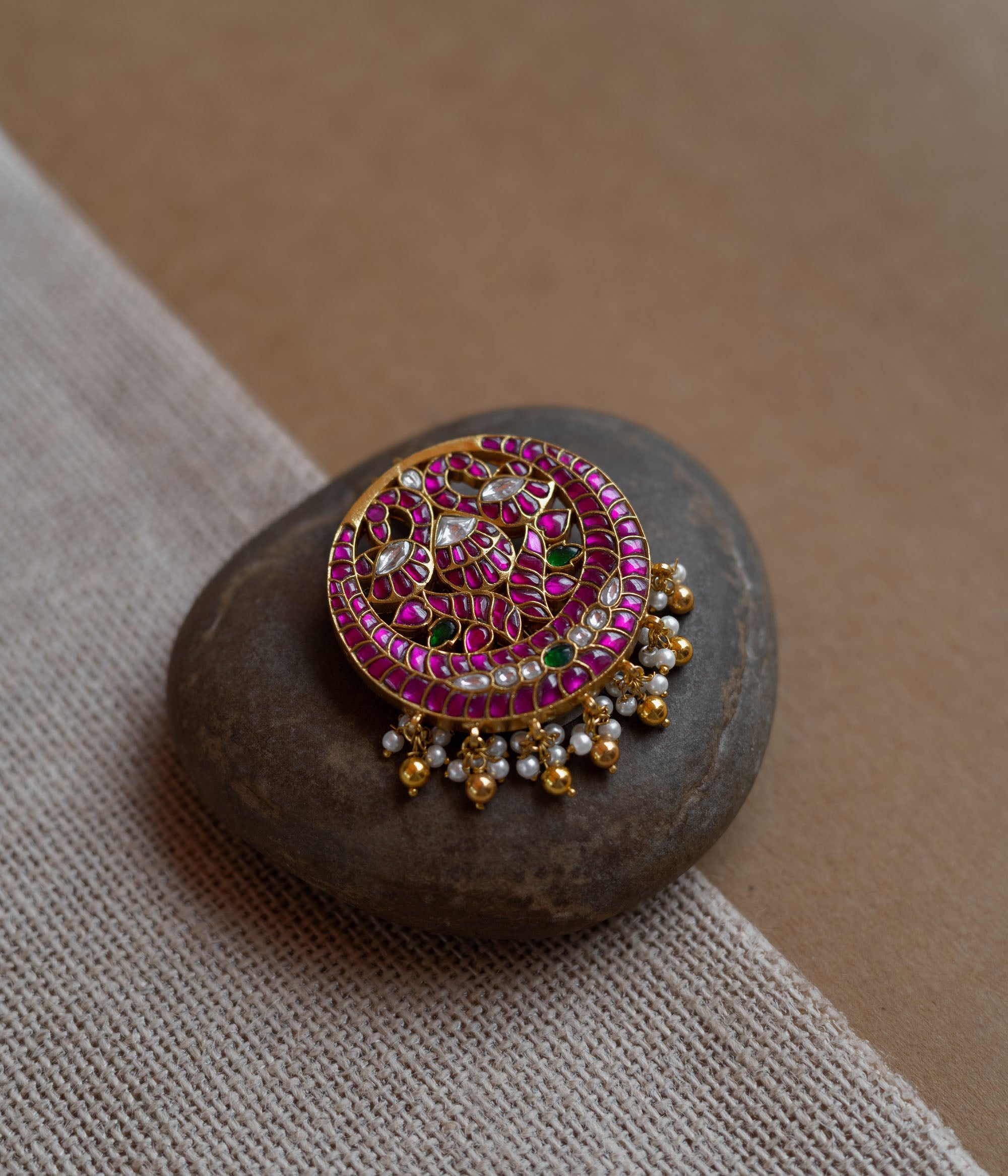 Close-up of a circular gold hairpin with a peacock motif. It's adorned with pink, green, and white Kundan stones and dangling pearls, resting on a stone.
