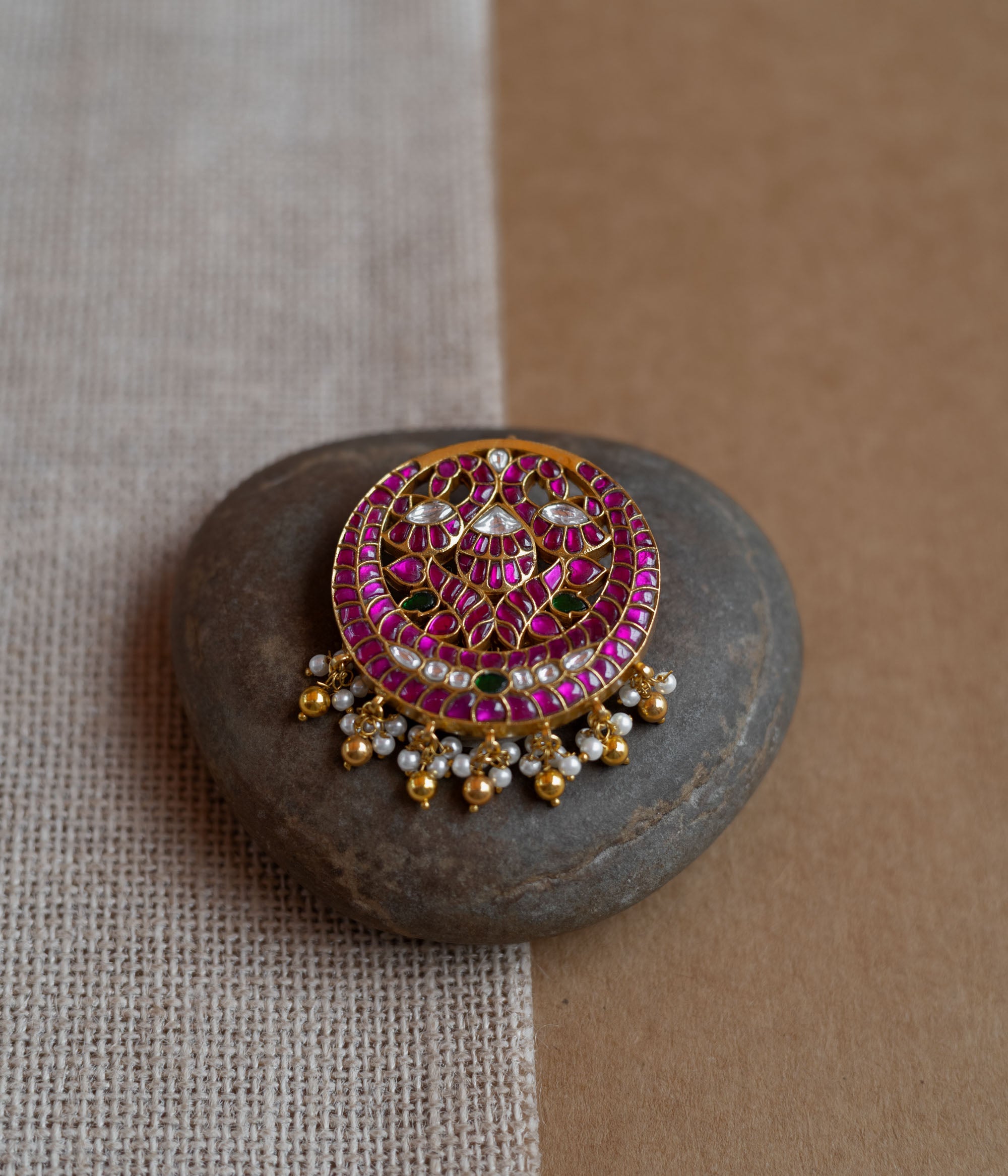 Close-up of a circular gold hairpin with a peacock motif. It's adorned with pink, green, and white Kundan stones and dangling pearls, resting on a stone.