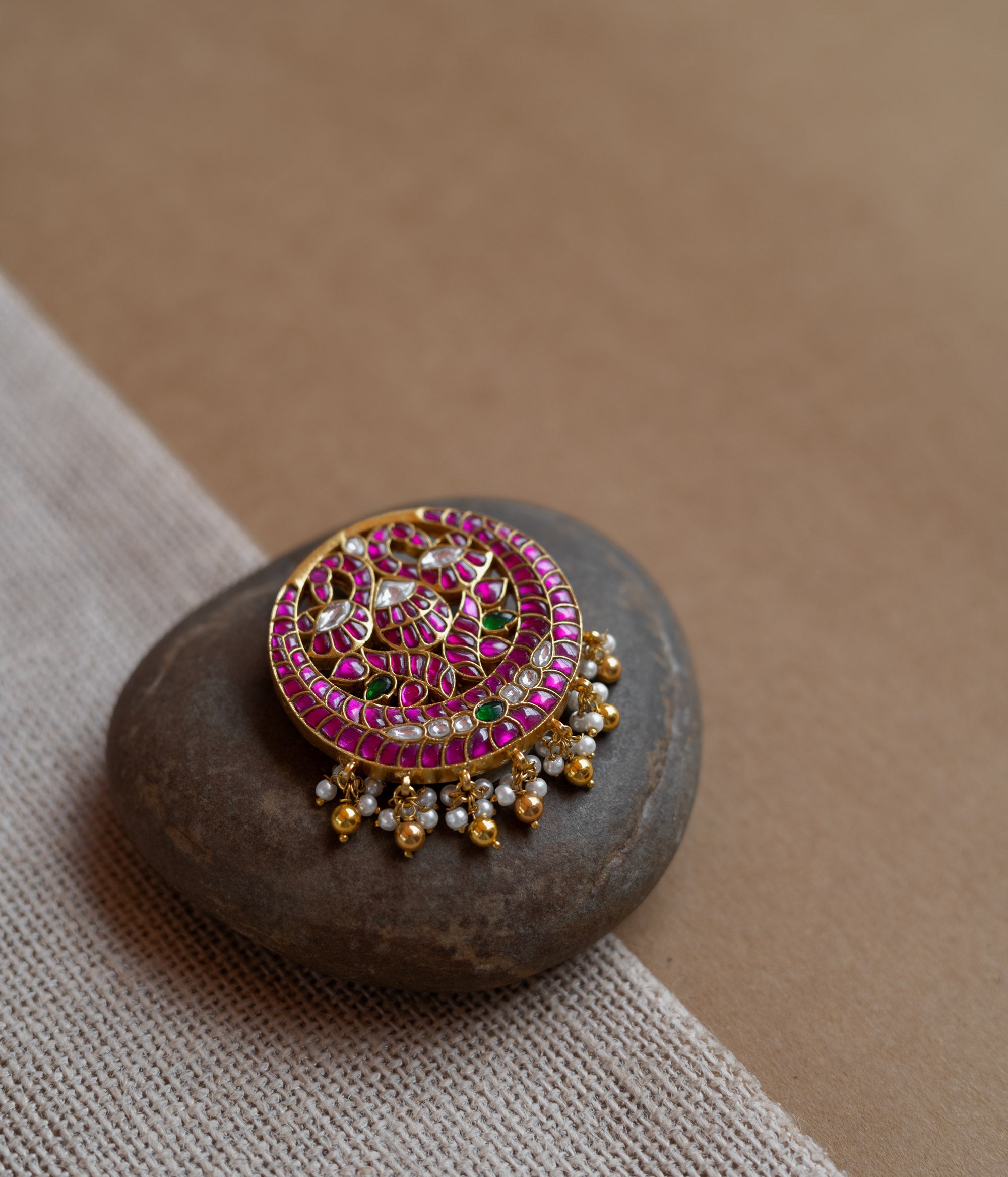 Close-up of a circular gold hairpin with a peacock motif. It's adorned with pink, green, and white Kundan stones and dangling pearls, resting on a stone.