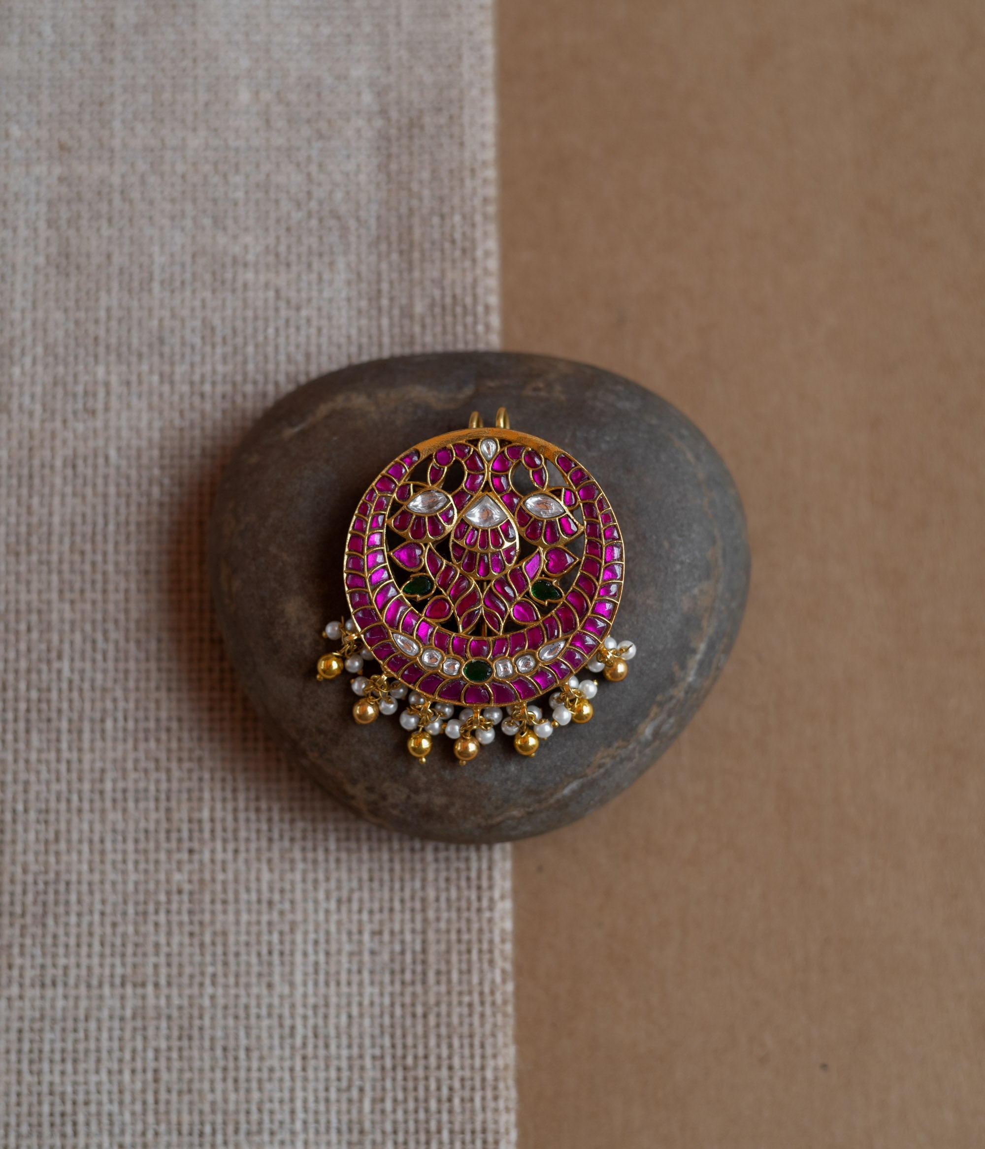 Close-up of a circular gold hairpin with a peacock motif. It's adorned with pink, green, and white Kundan stones and dangling pearls, resting on a stone.