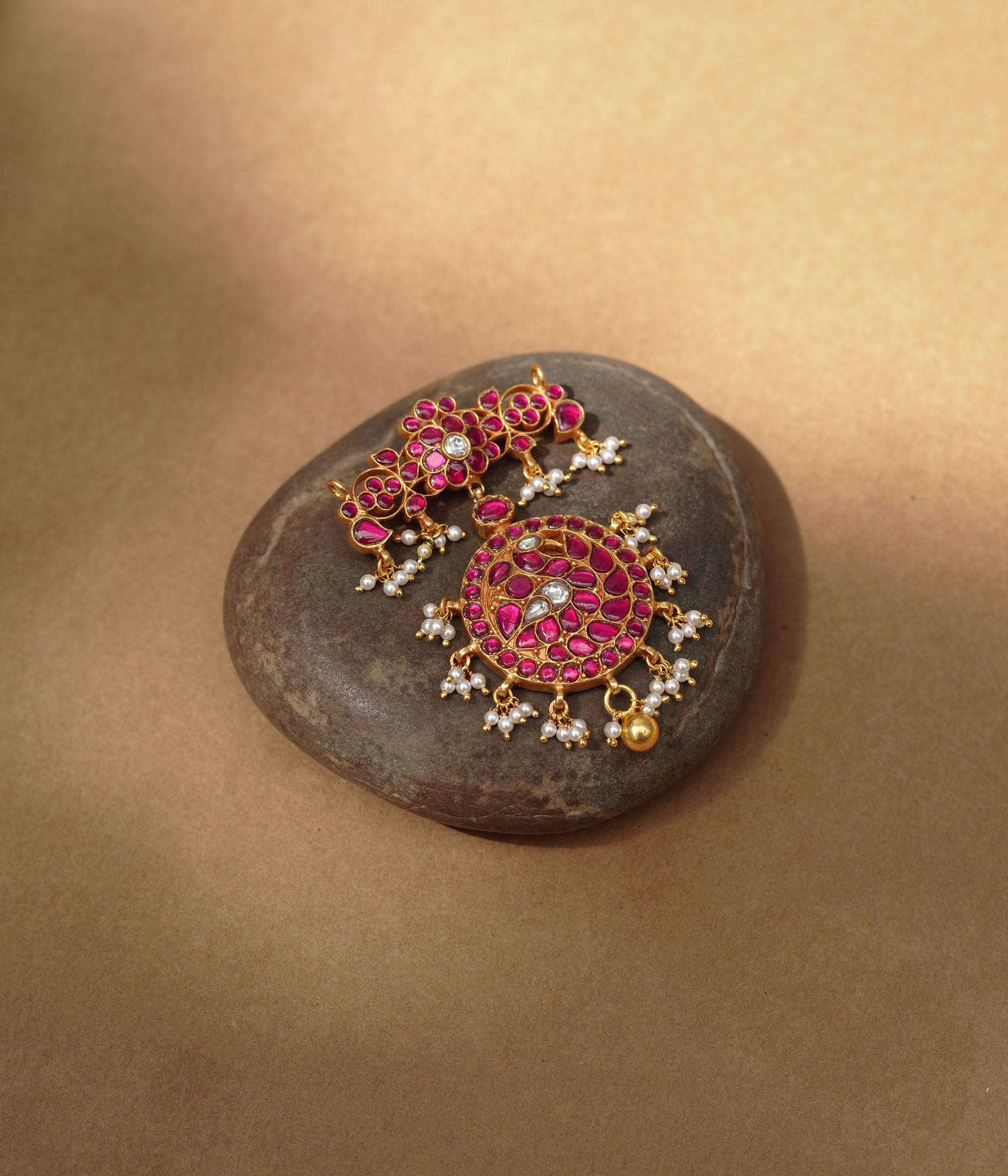 Close-up of a large gold Kundan pendant. It features a circular main section and a floral top piece, both adorned with red and white Kundan stones and dangling pearls.