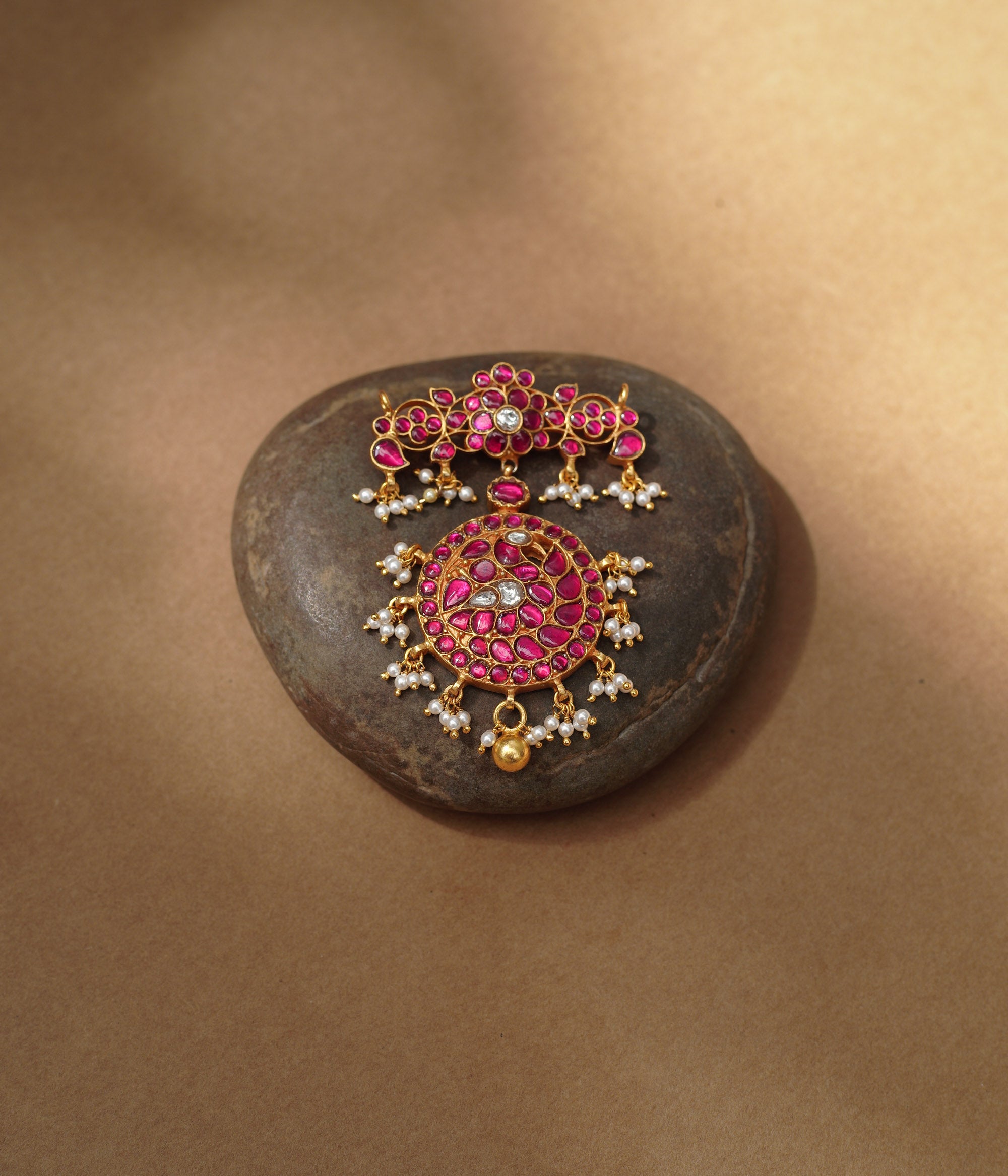 Close-up of a large gold Kundan pendant. It features a circular main section and a floral top piece, both adorned with red and white Kundan stones and dangling pearls.