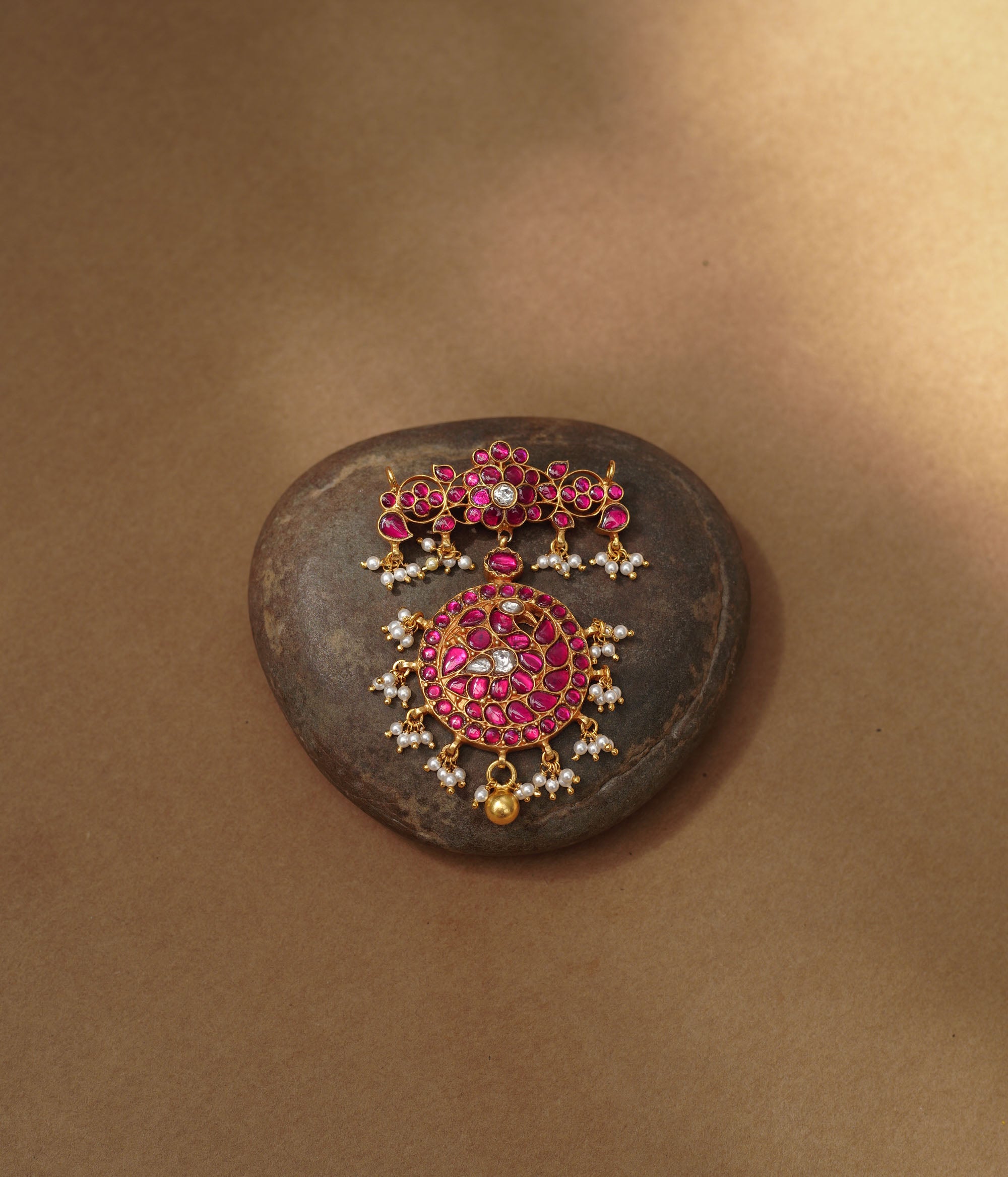 Close-up of a large gold Kundan pendant. It features a circular main section and a floral top piece, both adorned with red and white Kundan stones and dangling pearls.