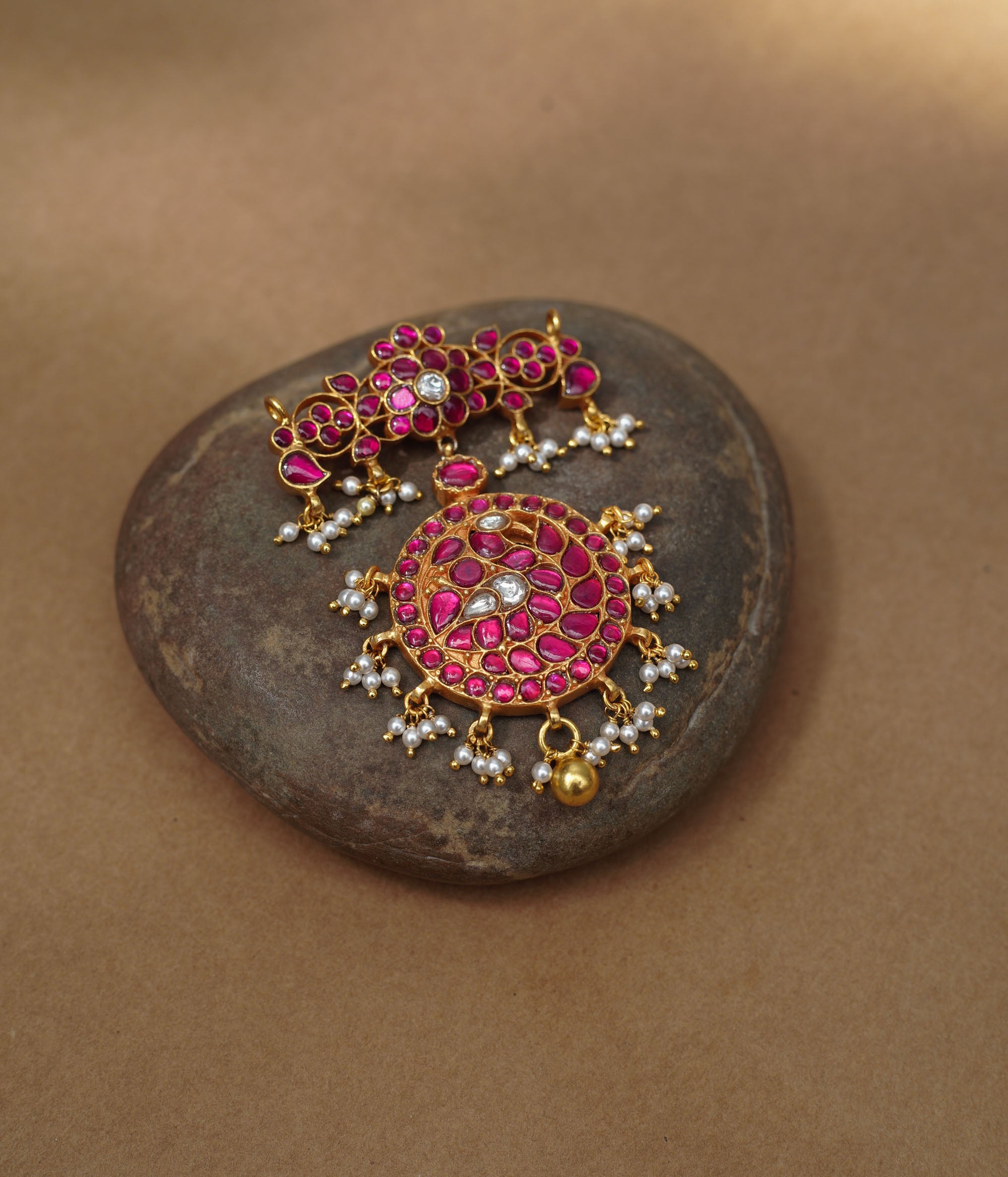 Close-up of a large gold Kundan pendant. It features a circular main section and a floral top piece, both adorned with red and white Kundan stones and dangling pearls.