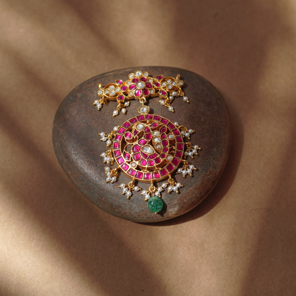 Close-up of a large gold Kundan pendant. It features a circular main section with red and white stones and dangling pearls, and a single green bead. A matching top piece is also visible.