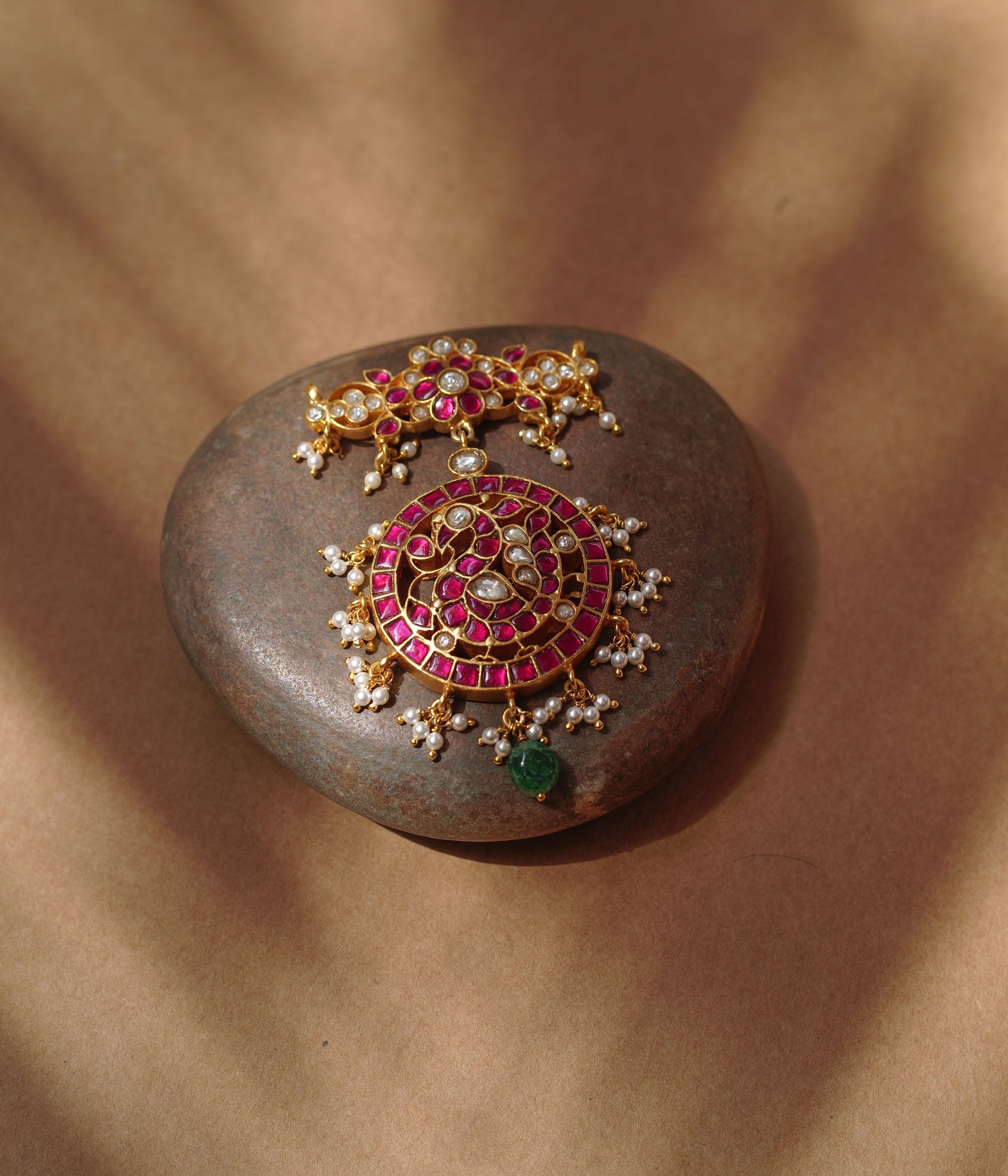 Close-up of a large gold Kundan pendant. It features a circular main section with red and white stones and dangling pearls, and a single green bead. A matching top piece is also visible.