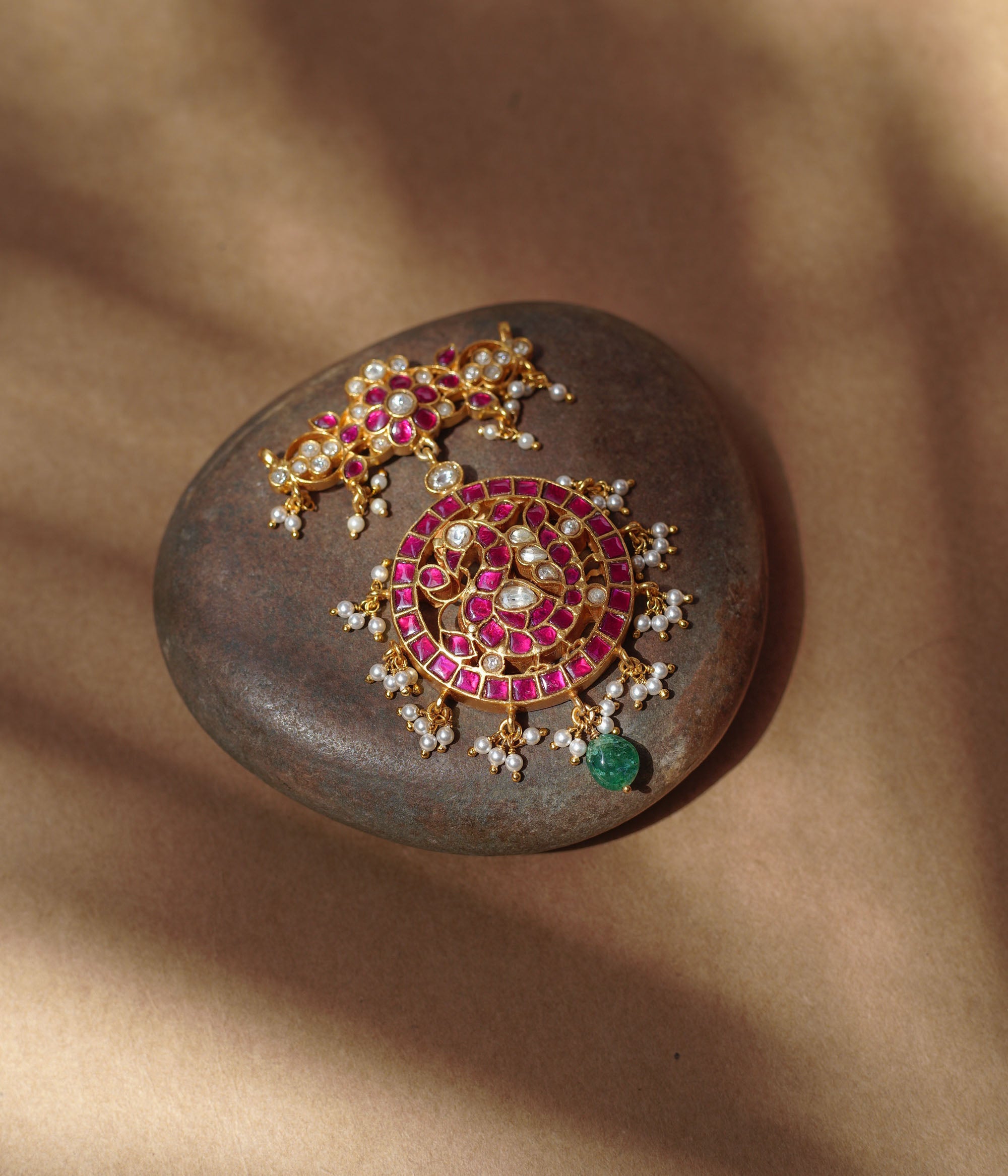 Close-up of a large gold Kundan pendant. It features a circular main section with red and white stones and dangling pearls, and a single green bead. A matching top piece is also visible.