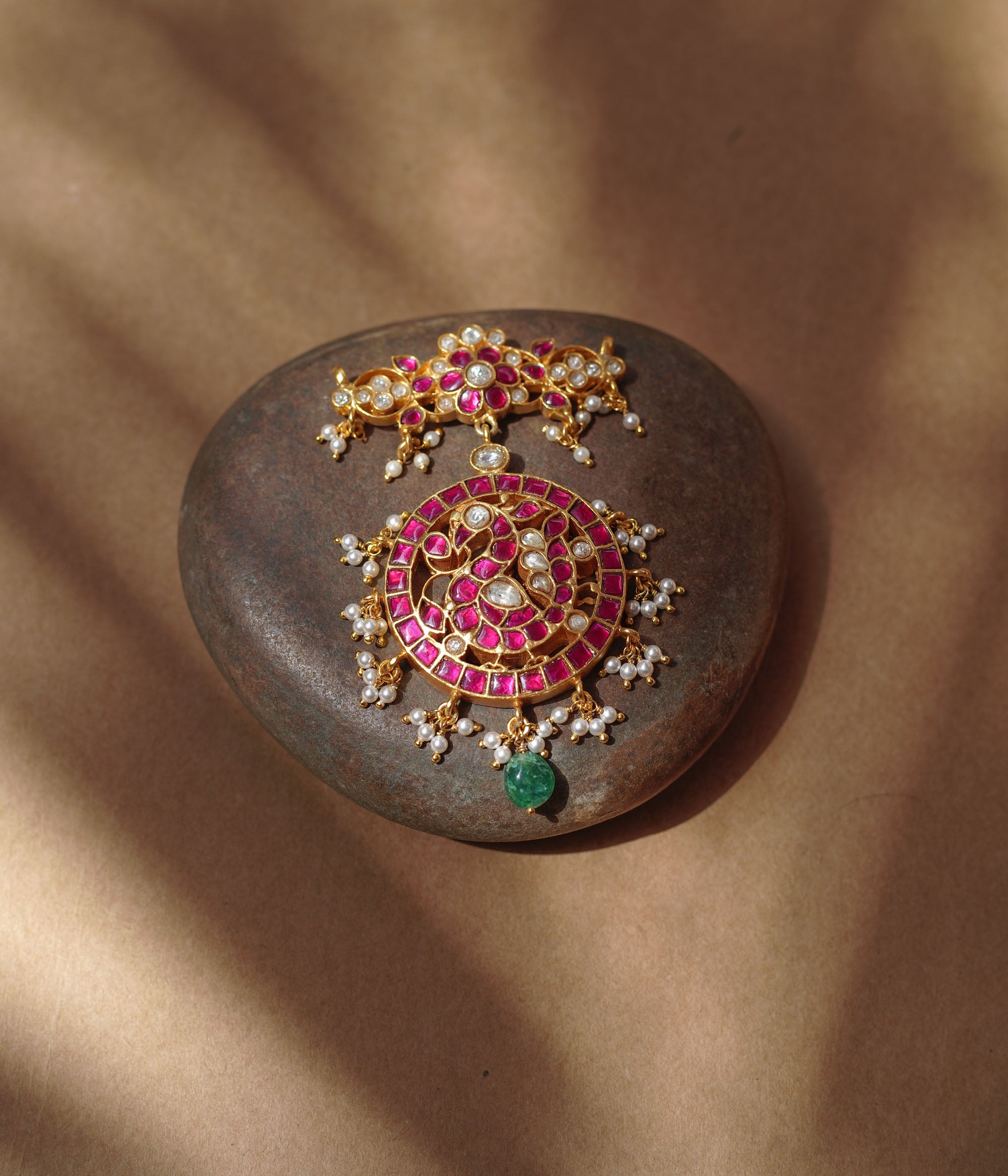 Close-up of a large gold Kundan pendant. It features a circular main section with red and white stones and dangling pearls, and a single green bead. A matching top piece is also visible.