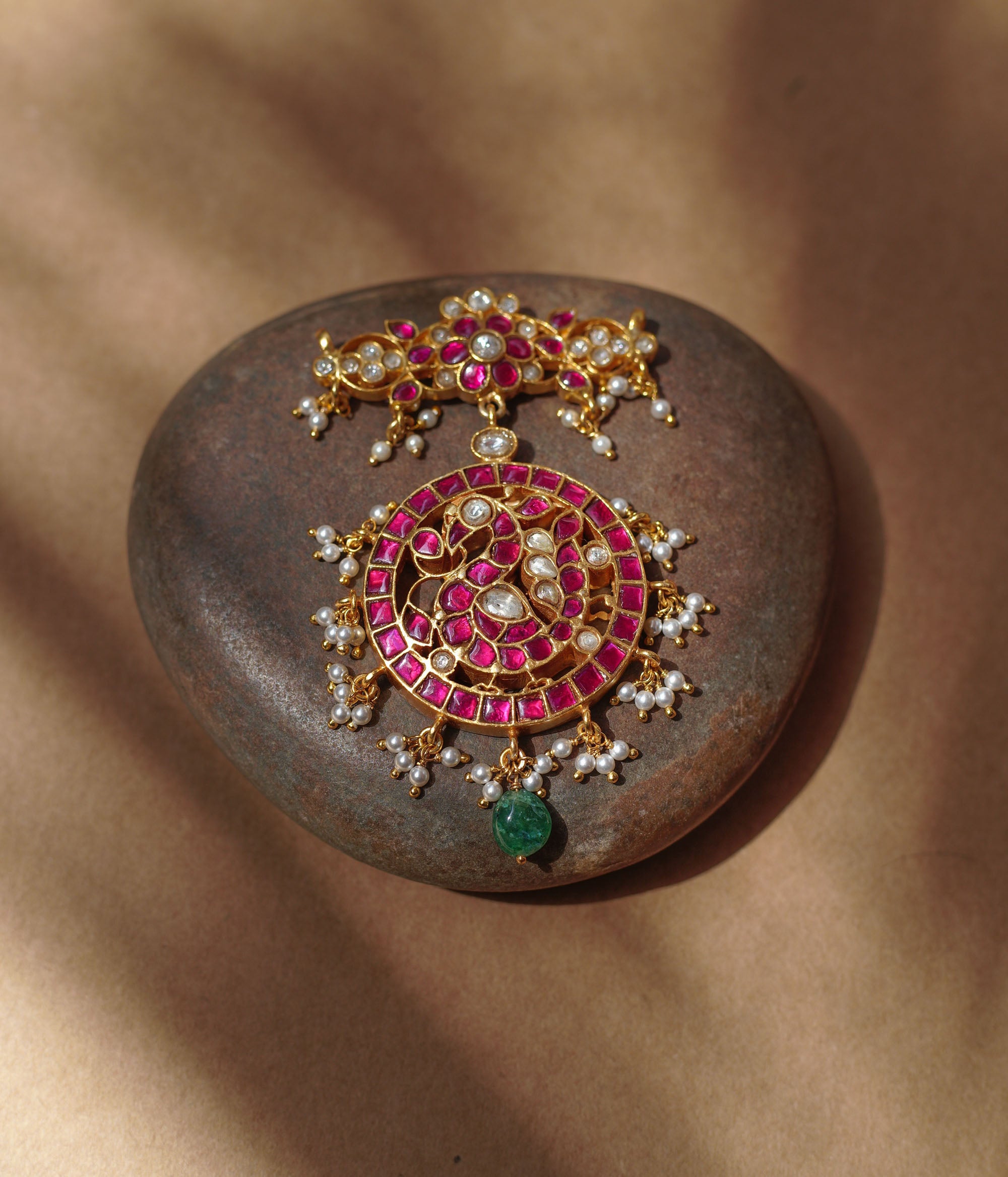 Close-up of a large gold Kundan pendant. It features a circular main section with red and white stones and dangling pearls, and a single green bead. A matching top piece is also visible.