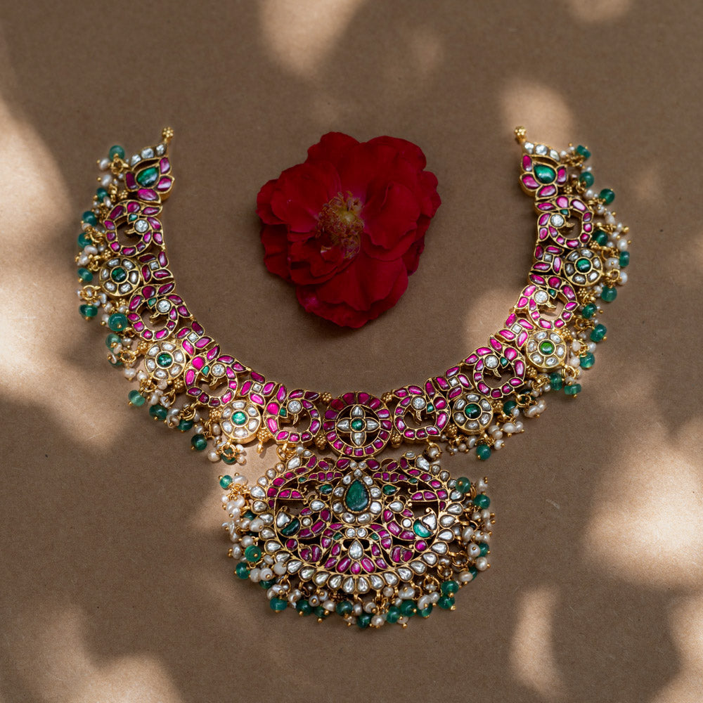 A handcrafted 22ct gold-plated 92.5 sterling silver Padmavati Kemp necklace, featuring red, green, and white Kundan stones, green beads, and pearls, with a broad, ornate design and a central pearl-clustered pendant, ideal for bridal wear and grand celebrations.