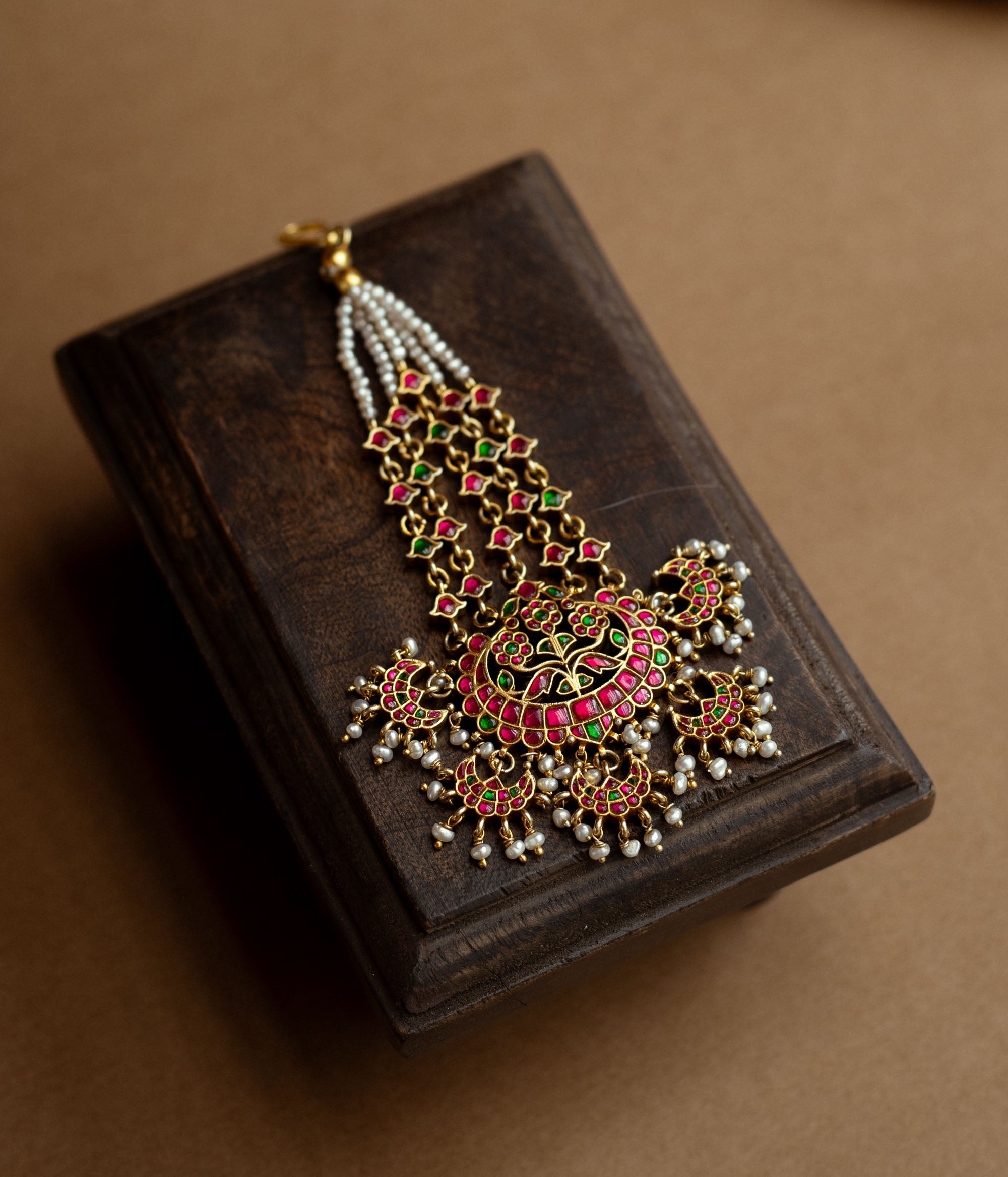 A floral tikka with green and red Kundan stones and dangling pearls.
