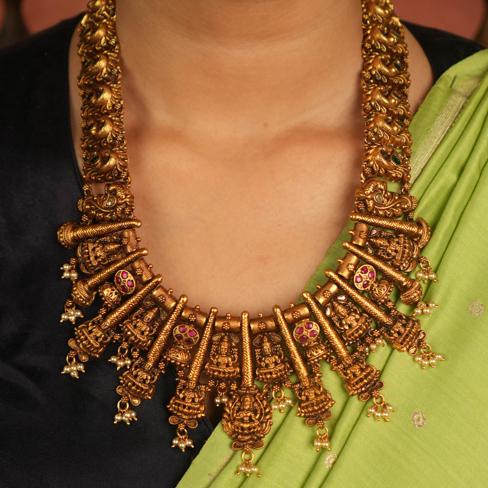 Temple whispers reborn! This antique gold-plated necklace features a continuous chain of temple jewelry adorned with intricate Goddess Lakshmi motifs. Red and green Kundan stones bloom with divine grace, while delicate pearl droplets add a touch of ethereal charm. A timeless treasure that embodies heritage, devotion, and exquisite artistry.