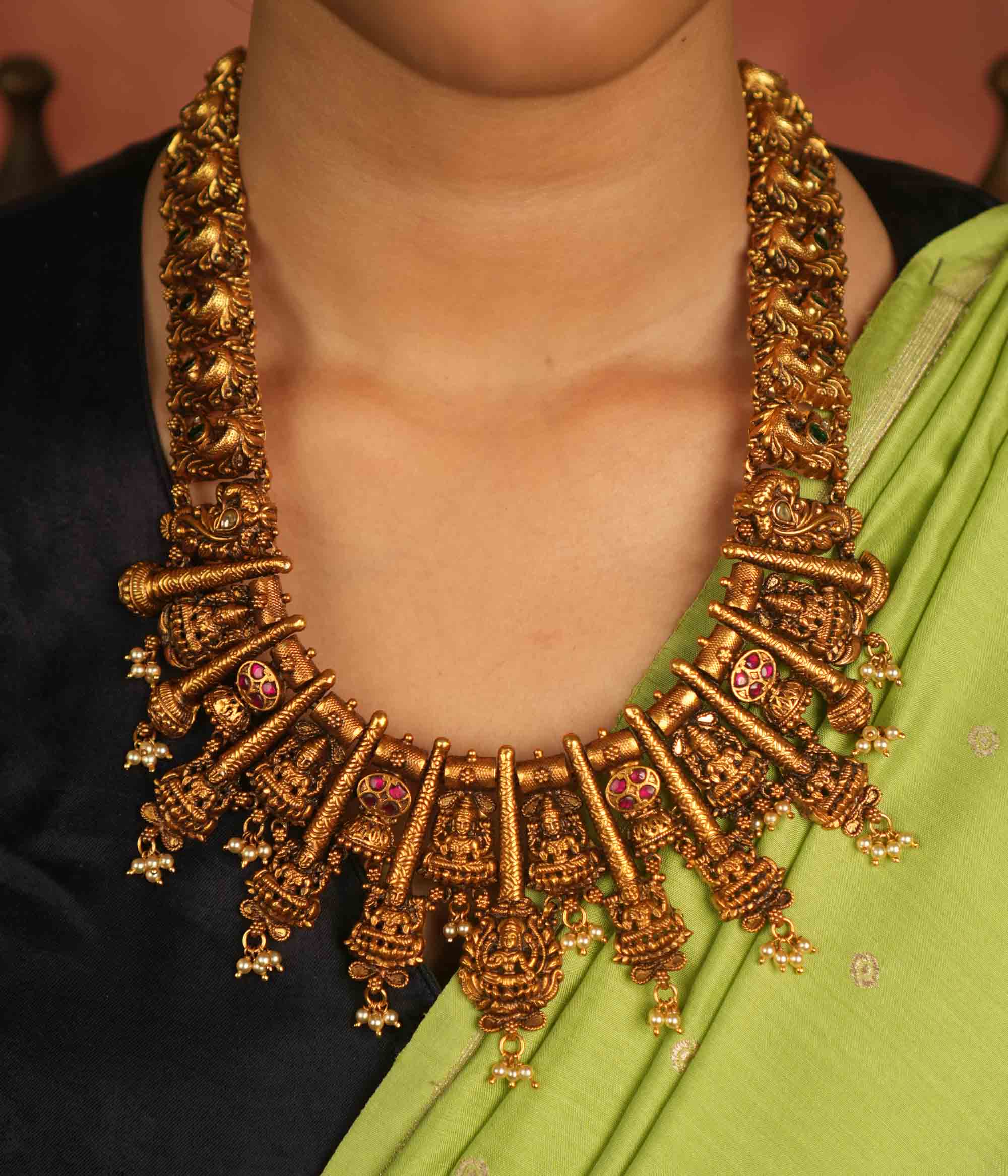 Temple whispers reborn! This antique gold-plated necklace features a continuous chain of temple jewelry adorned with intricate Goddess Lakshmi motifs. Red and green Kundan stones bloom with divine grace, while delicate pearl droplets add a touch of ethereal charm. A timeless treasure that embodies heritage, devotion, and exquisite artistry.