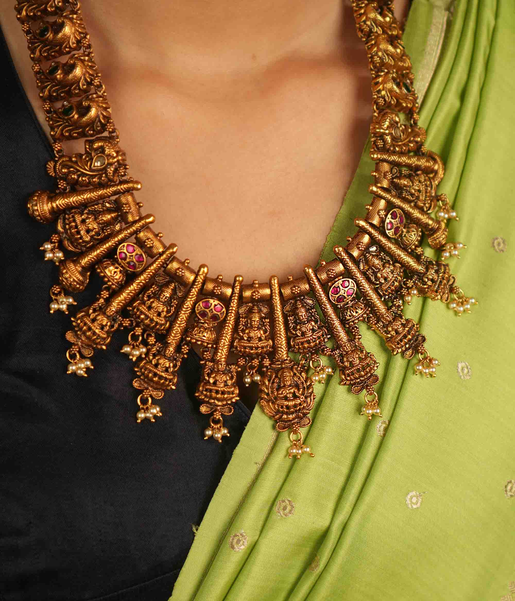 Temple whispers reborn! This antique gold-plated necklace features a continuous chain of temple jewelry adorned with intricate Goddess Lakshmi motifs. Red and green Kundan stones bloom with divine grace, while delicate pearl droplets add a touch of ethereal charm. A timeless treasure that embodies heritage, devotion, and exquisite artistry.