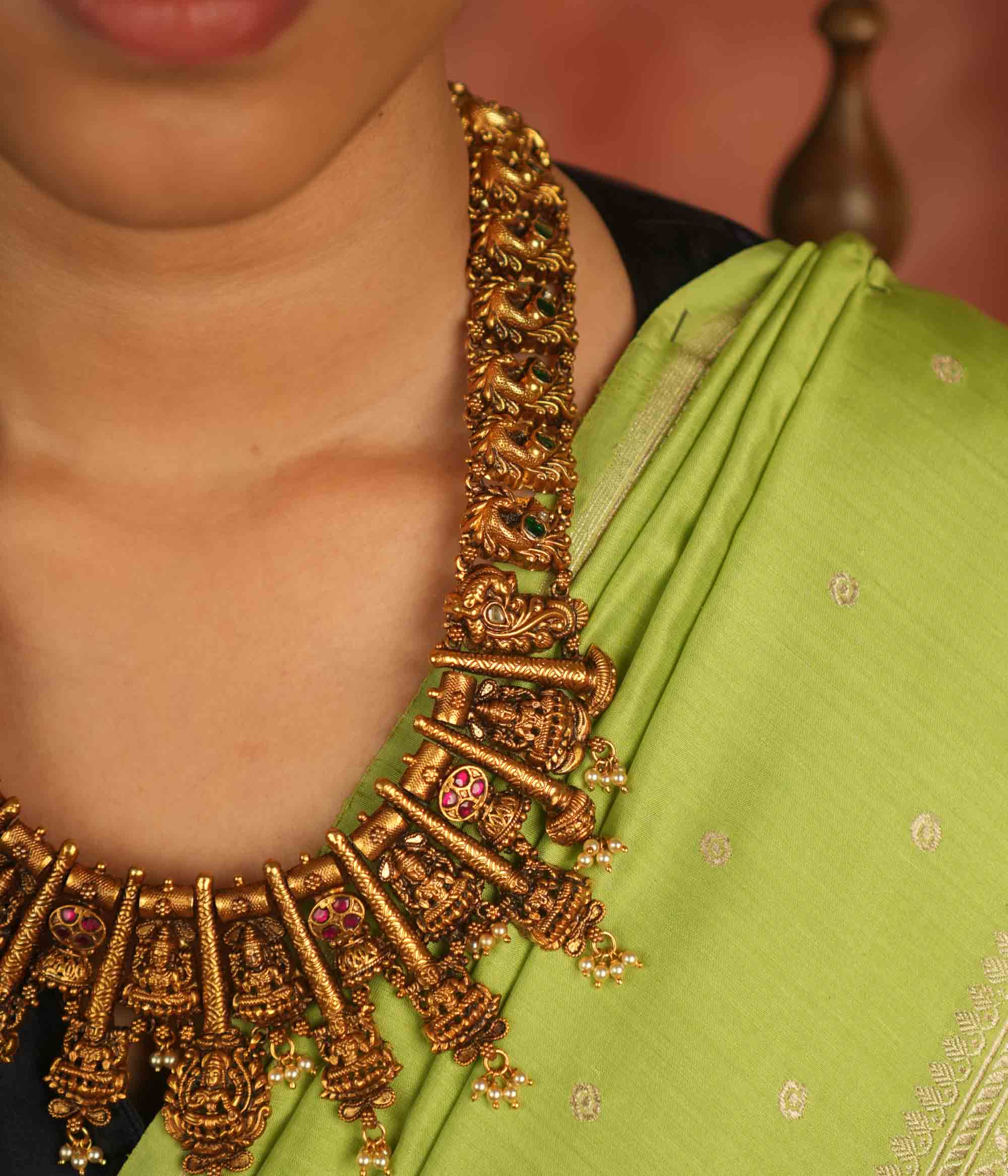 Temple whispers reborn! This antique gold-plated necklace features a continuous chain of temple jewelry adorned with intricate Goddess Lakshmi motifs. Red and green Kundan stones bloom with divine grace, while delicate pearl droplets add a touch of ethereal charm. A timeless treasure that embodies heritage, devotion, and exquisite artistry.