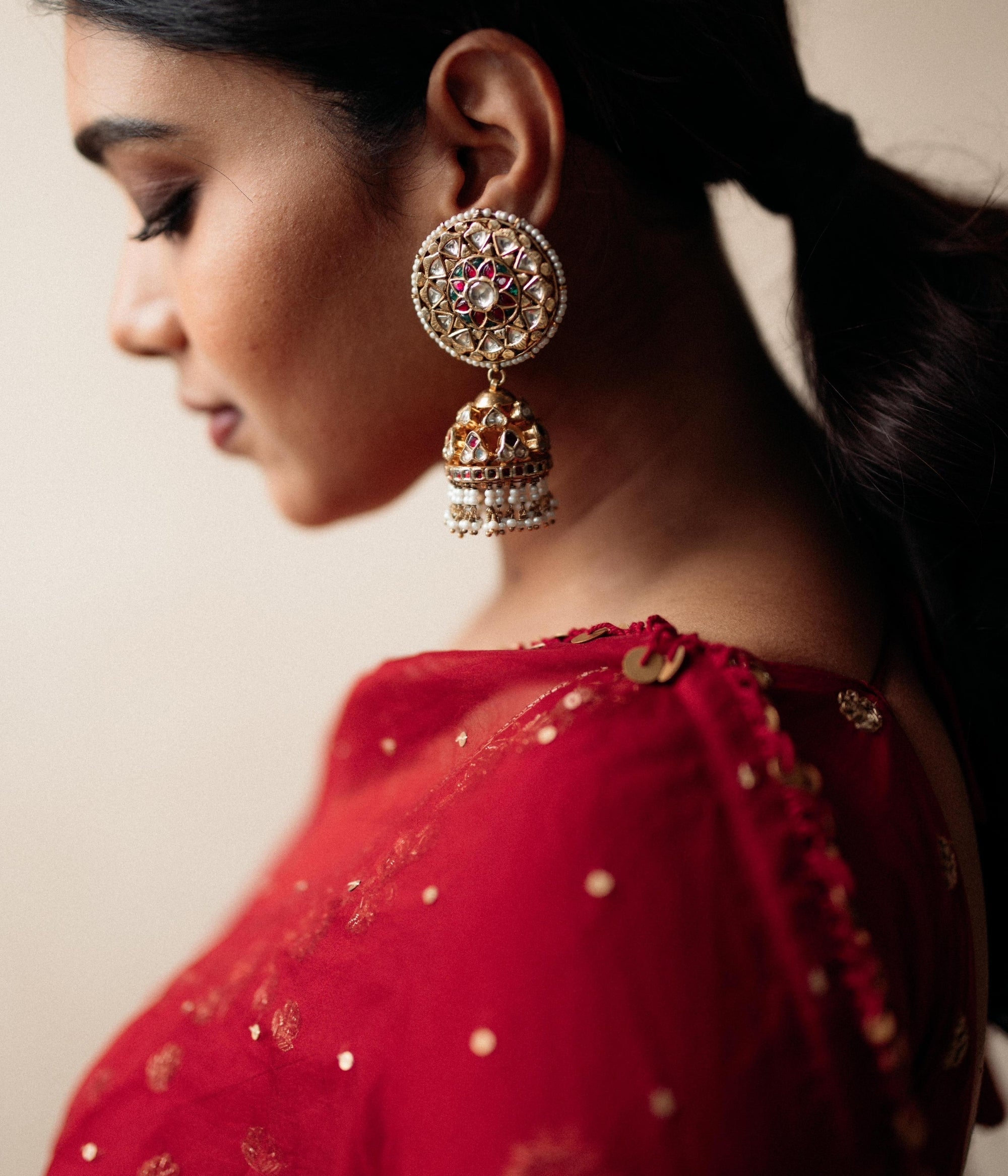 Large gold jhumka earrings with white sapphires, rubies, and pearls, on a woman wearing a red sari.