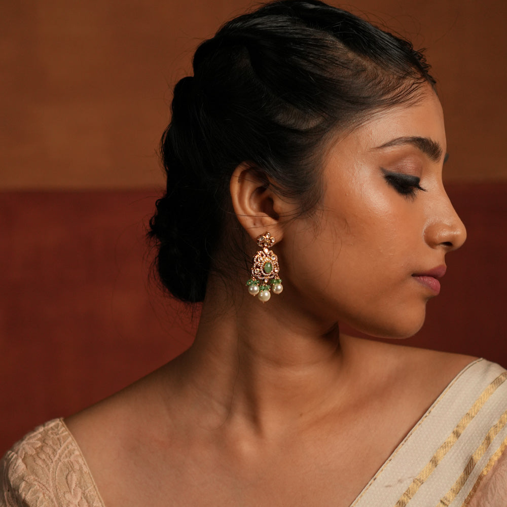 Exquisite gold-plated silver dangler earrings featuring a floral stud with a dazzling Polki stone at the center, encased in shimmering CZ stones. The elegant drop showcases a vibrant green gemstone in the middle, surrounded by Polki and CZ stones, forming a graceful floral design. Delicate white pearl and green bead drops enhance its charm, adding a touch of sophistication and tradition to any look.