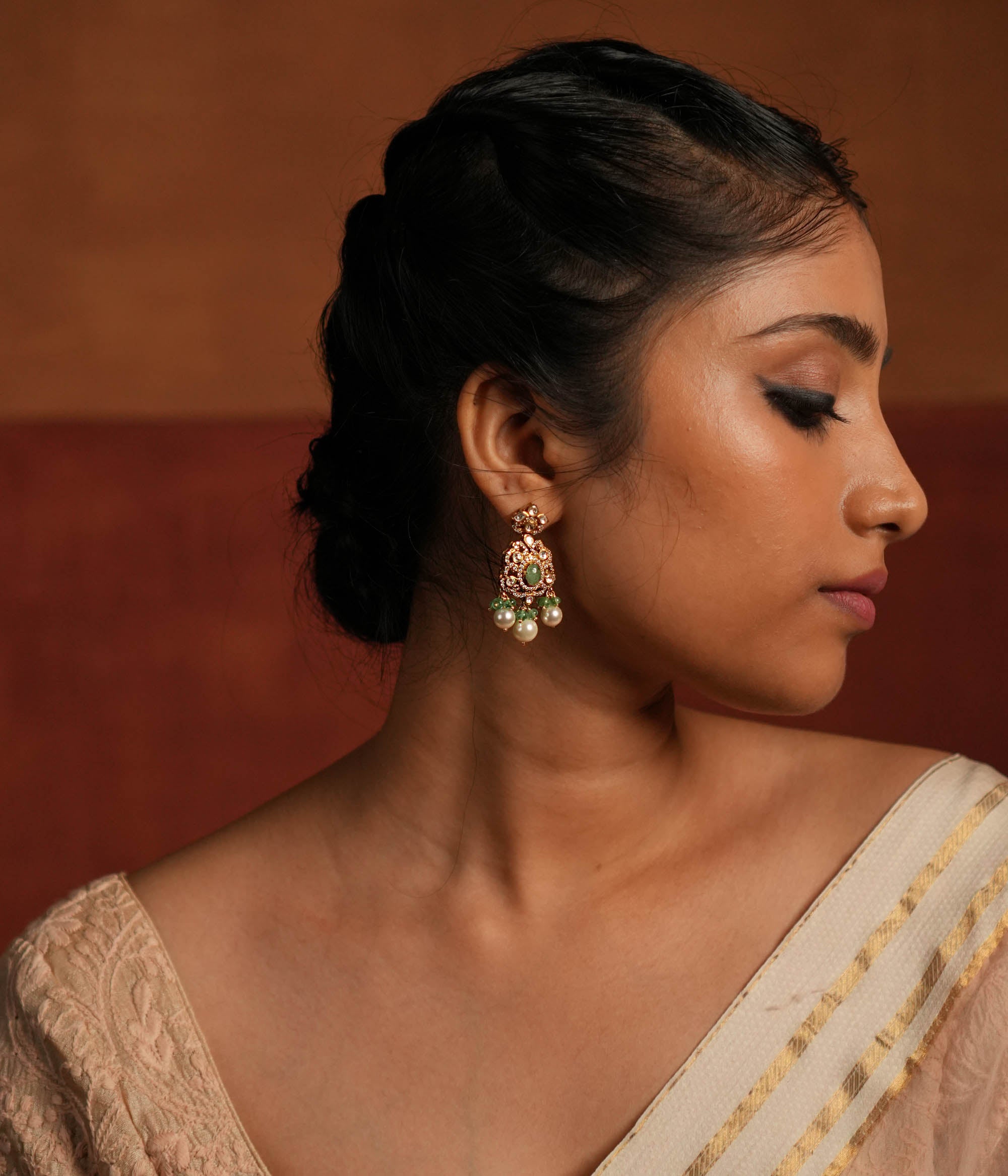 Exquisite gold-plated silver dangler earrings featuring a floral stud with a dazzling Polki stone at the center, encased in shimmering CZ stones. The elegant drop showcases a vibrant green gemstone in the middle, surrounded by Polki and CZ stones, forming a graceful floral design. Delicate white pearl and green bead drops enhance its charm, adding a touch of sophistication and tradition to any look.