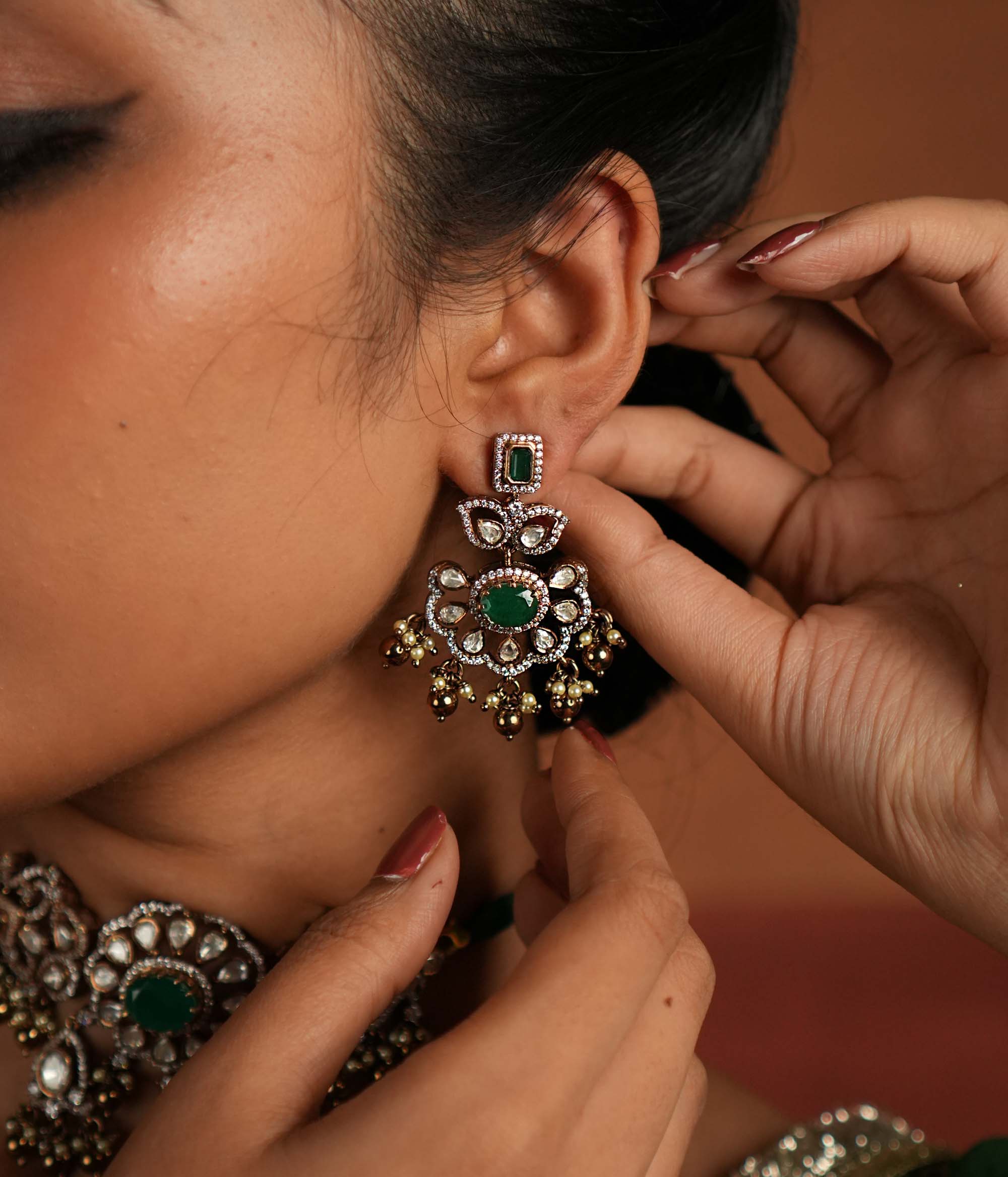 Victorian elegance reborn! These gold-plated silver Chandbali earrings showcase a radiant green gemstone at the center, encircled by shimmering polki and CZ stones. Graceful pearl drops with golden beads add a touch of charm, evoking the allure of a forgotten era. A secret garden of beauty, crafted to adorn the modern woman.