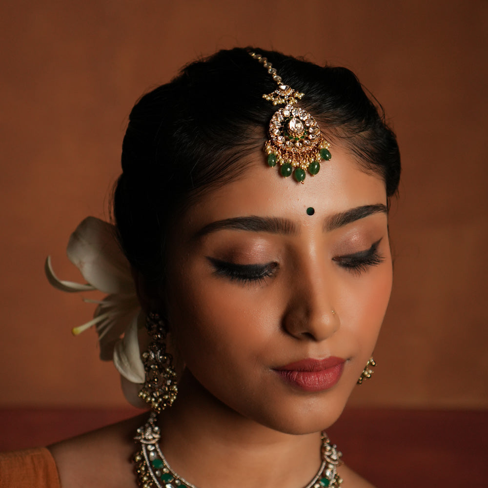 Victorian elegance reborn! This gold-plated silver maangtika features a stunning floral pendant with a central moissanite polki stone, encircled by smaller polkis and CZ stones. Graceful drops of green beads and pearls cascade beautifully, evoking whispers of forgotten lore. A timeless crown of sophistication, reimagined for the modern bride.