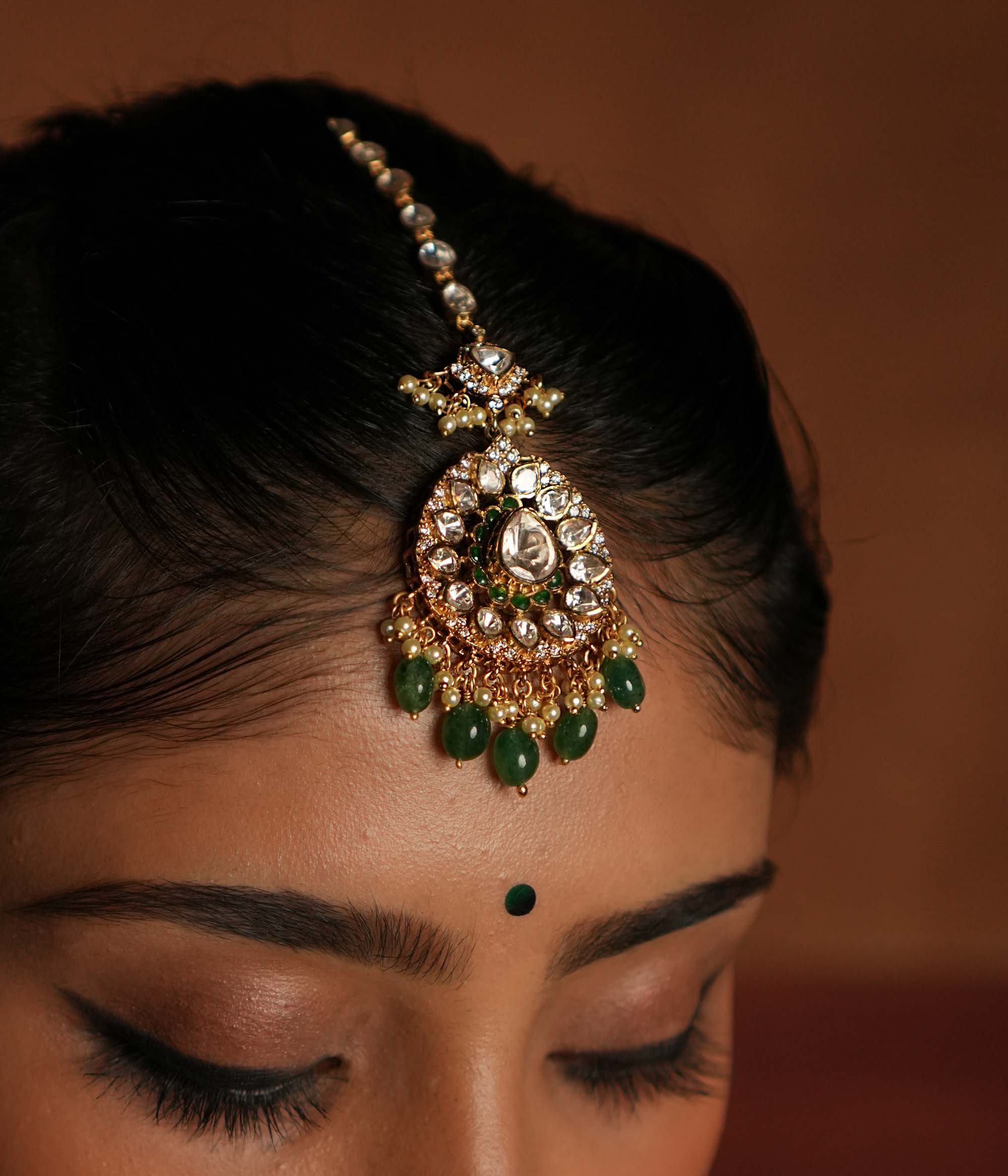 Victorian elegance reborn! This gold-plated silver maangtika features a stunning floral pendant with a central moissanite polki stone, encircled by smaller polkis and CZ stones. Graceful drops of green beads and pearls cascade beautifully, evoking whispers of forgotten lore. A timeless crown of sophistication, reimagined for the modern bride.
